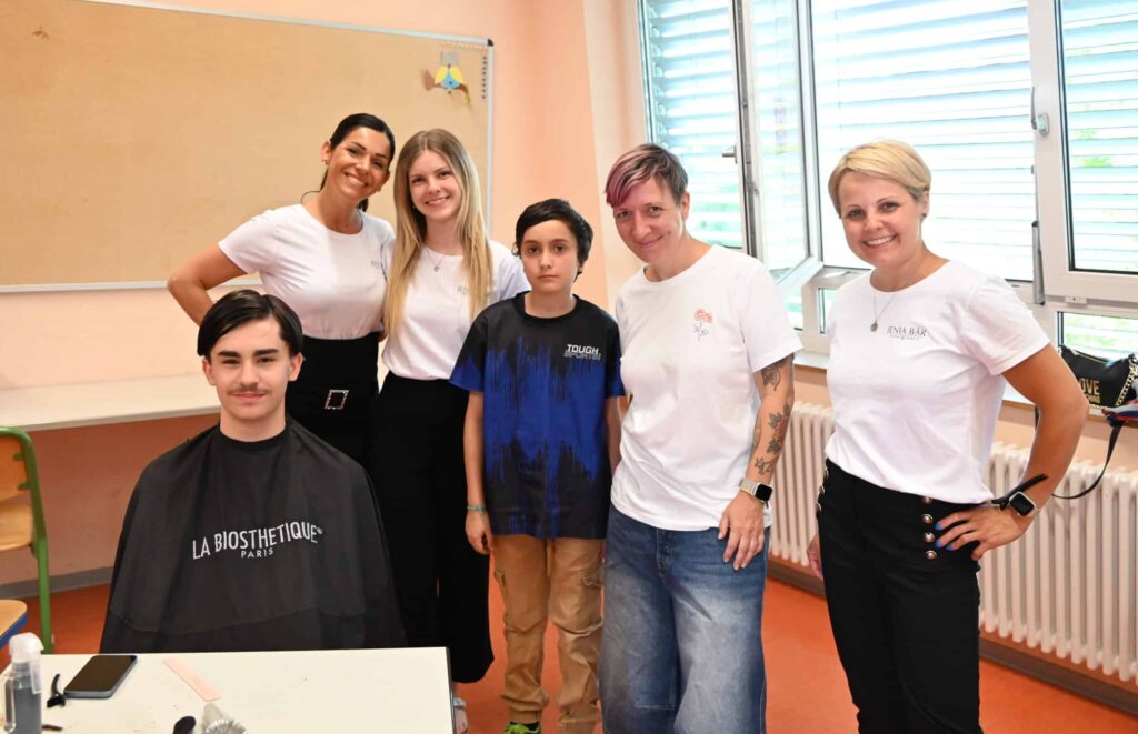 Eine Gruppe von sechs Personen. Ein Junge sitzt, er trägt einen Frisörumhang. Alle anderen stehen. Die vier Frauen tragen weiße T-Shirts, der Junge in Ihrer Mitte trägt ein schwarzes T-Shirt. Die lachen in die Kamera.