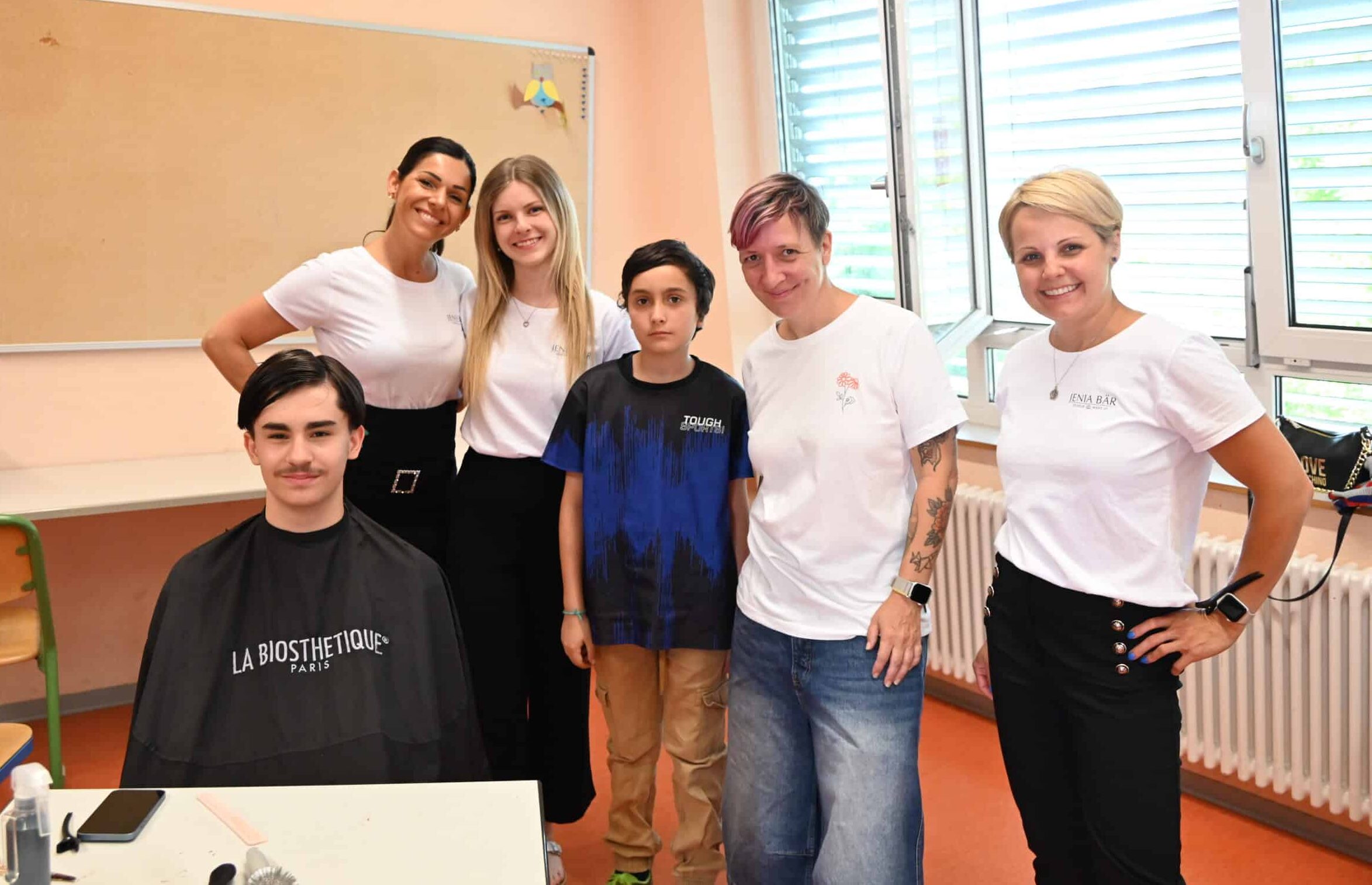 Eine Gruppe von sechs Personen. Ein Junge sitzt, er trägt einen Frisörumhang. Alle anderen stehen. Die vier Frauen tragen weiße T-Shirts, der Junge in Ihrer Mitte trägt ein schwarzes T-Shirt. Die lachen in die Kamera.