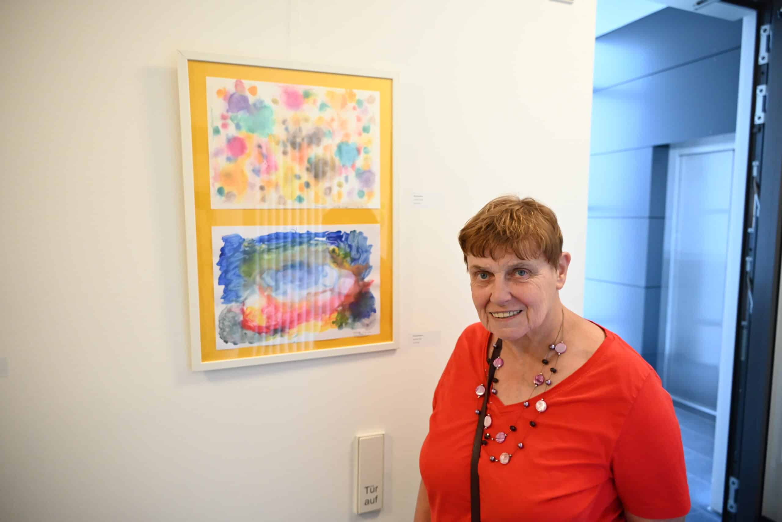 Eine Frau im roten T-Shirt steht vor einer weißen Wand, an der ein Bild hängt. Sie lächelt in die Kamera.