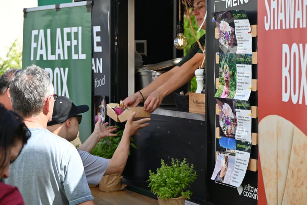 Eine Frau reicht durch das Fenster des Foodtrucks eine Box mit Essen. Eine andere Person nimmt das Essen entgegen.