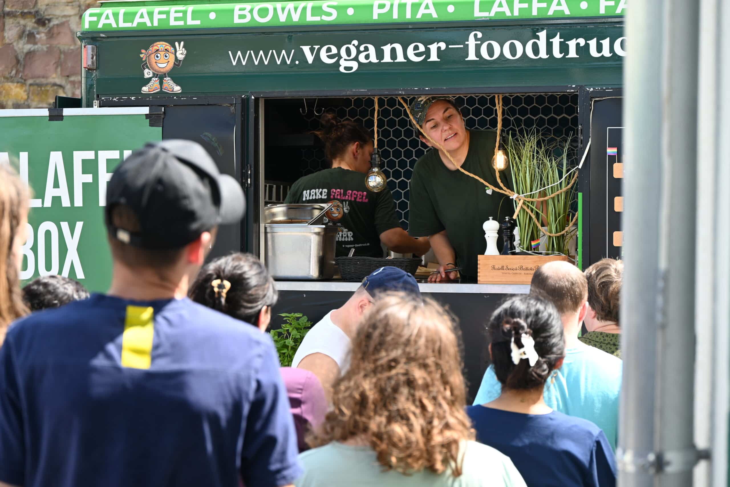 Quelle: BLV Eine Frau schaut fragend aus dem Fenster des Foodtrucks. Sie nimmt die neuen Essensbestellungen auf. Viele Menschen warten vor dem Foodtruck.