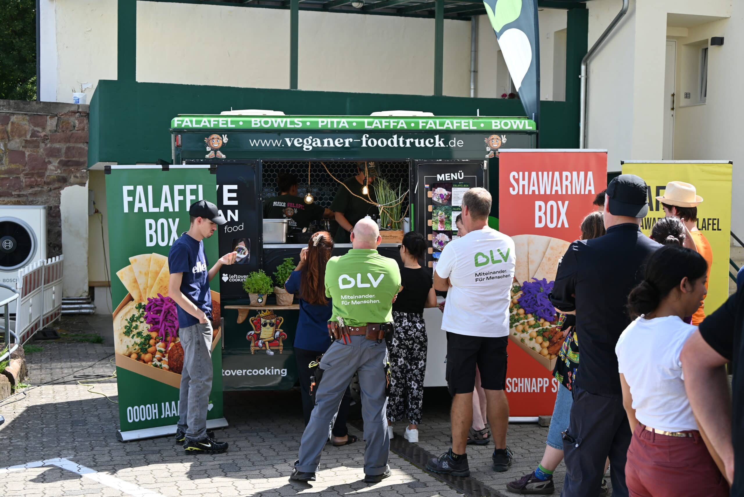 Quelle: BLV Eine lange Schlange Menschen steht vor einem Foodtruck an und wartet darauf, Essen zu bestellen. Durch ein Fenster sieht man im Innern des Foodtrucks zwei Frauen, wie sie Essen zubereiten. In der Gruppe vor dem Foodtruck sind auch zwei Mitarbeiter des BLV - einer im grünen, der andere im weißen T-Shirt.