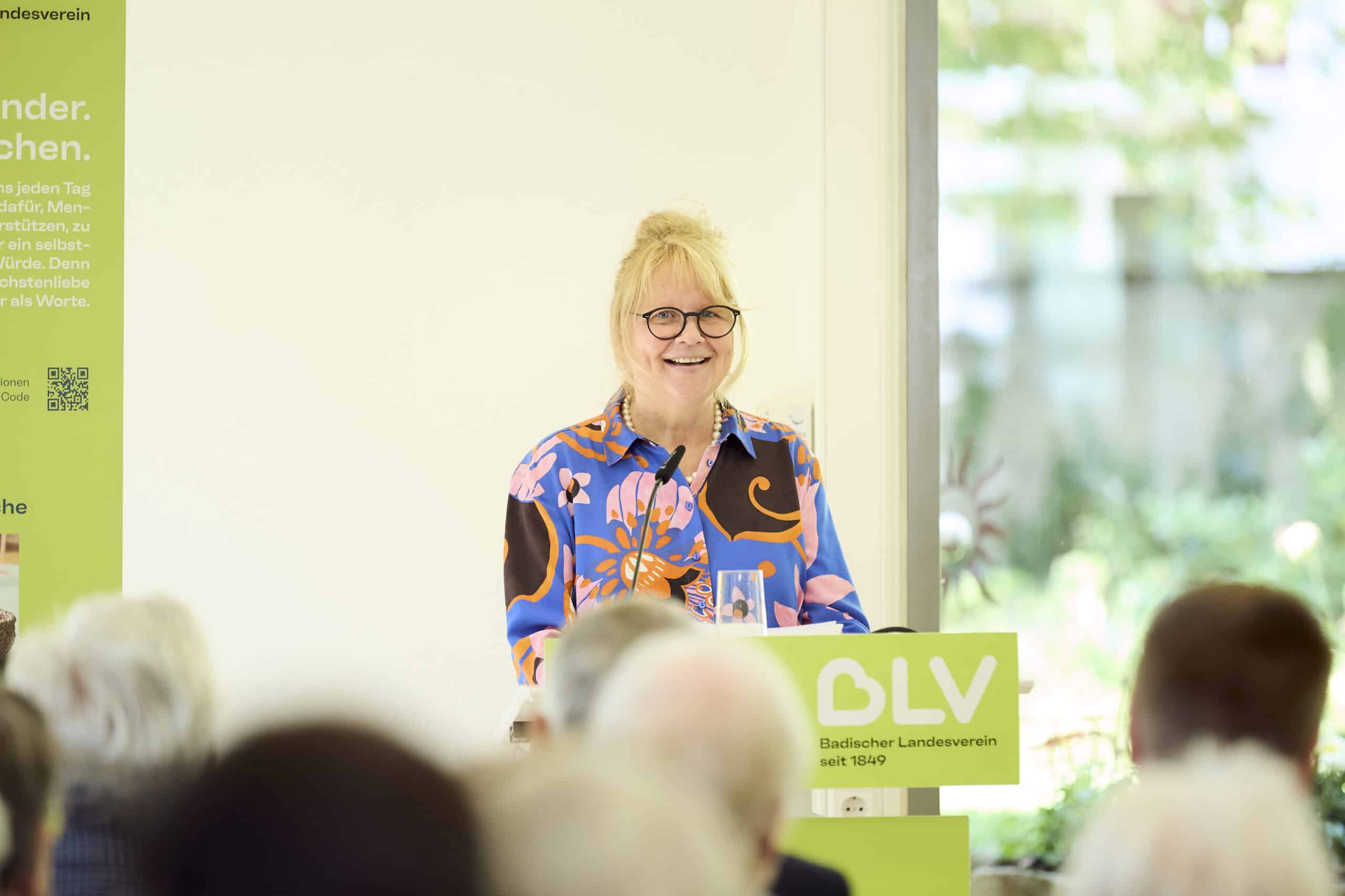 BLV-Vorstand Christine Jung-Weyand steht an einem Pult und hält eine Rede. Sie trägt eine bunt gemusterte Bluse und lächelt in die Kamera.