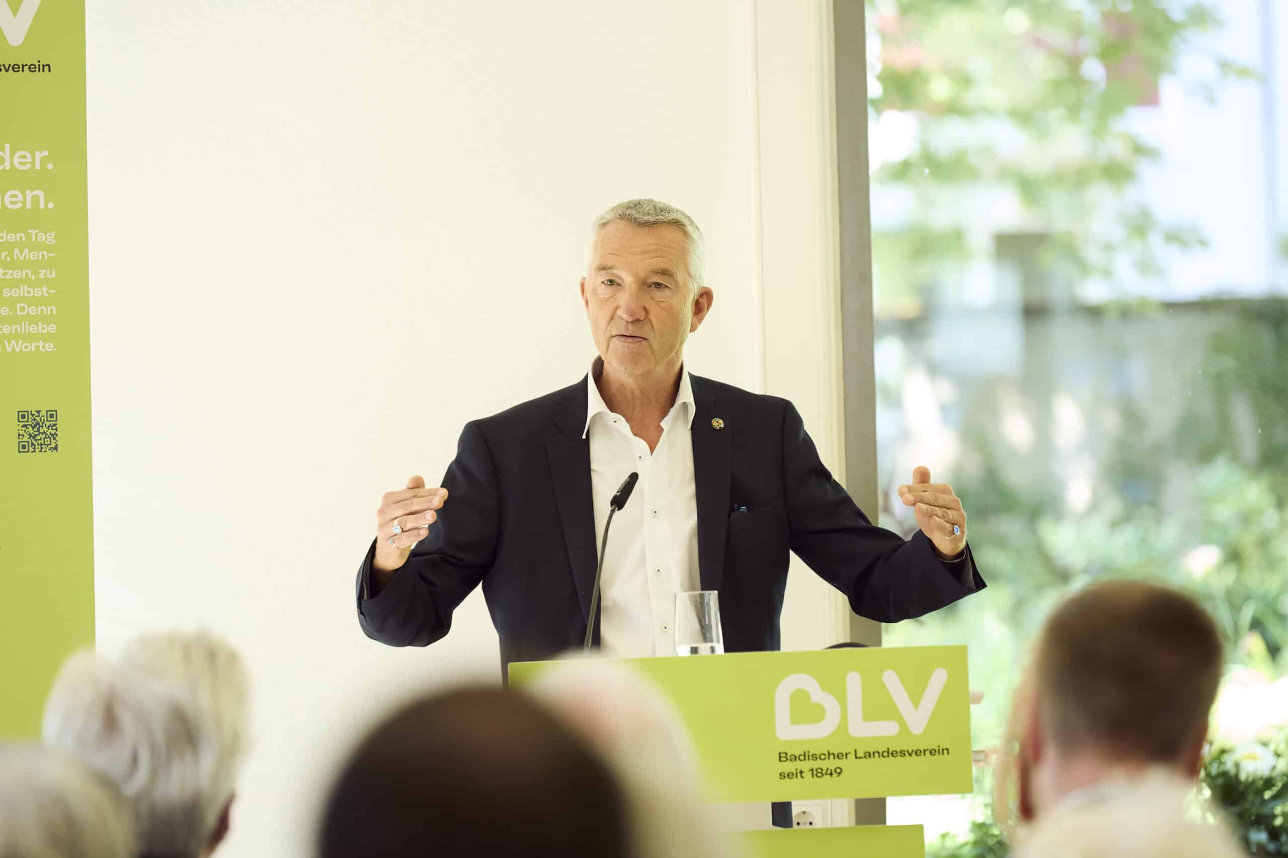 Karlsruhes Bürgermeister Dr. Martin Lenz steht an einem Pult und hält eine Rede. Er trägt einen dunkelblauen Anzug und ein weißes Hemd. Er habt beide Arme leicht an, wie um das Gesagte zu unterstreichen.