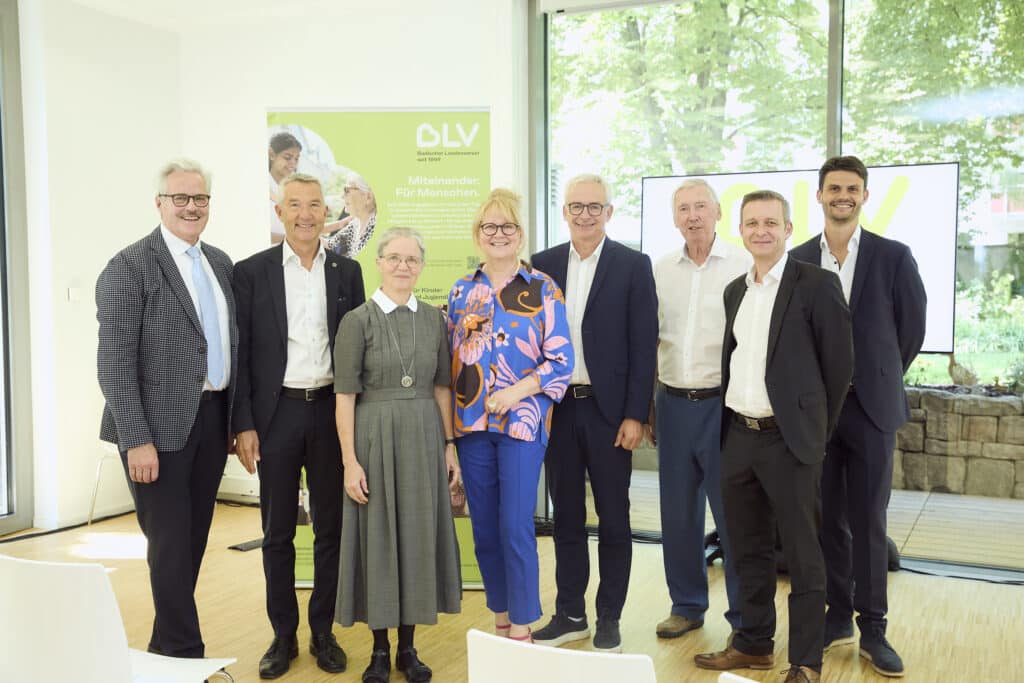 Eine Gruppe Menschen steht nebeneinander und lächelt in die Kamera. Sie tragen festliche Kleidung. Im Hintergrund sieht man einen Aufsteller des BLV und blickt durch große Fenster auf die Bäume im Garten des Friedensheims. Von links nach rechts sind zu sehen: Willy Kolarsch (Geschäftsführung GIG mbH), Dr. Martin Lenz (Bürgermeister Stadt Karlsruhe), Oberin Schwester Hildegund Fieg (Evangelisches Diakonissenhaus Bethlehem), Christine Jung-Weyand (Vorstand BLV), Dr. Christoph Schnaudigel (Landrat Landkreis Karlsruhe), Prof. Axel Göhringer (Verwaltungsratsvorsitzender BLV), Frank Derix (BWKG Baden-Württembergische Krankenhausgesellschaft e.V.), Pablo Müller Camarena (Geschäftsführung ACE Ingenieurconsult GmbH & Co. KG.)