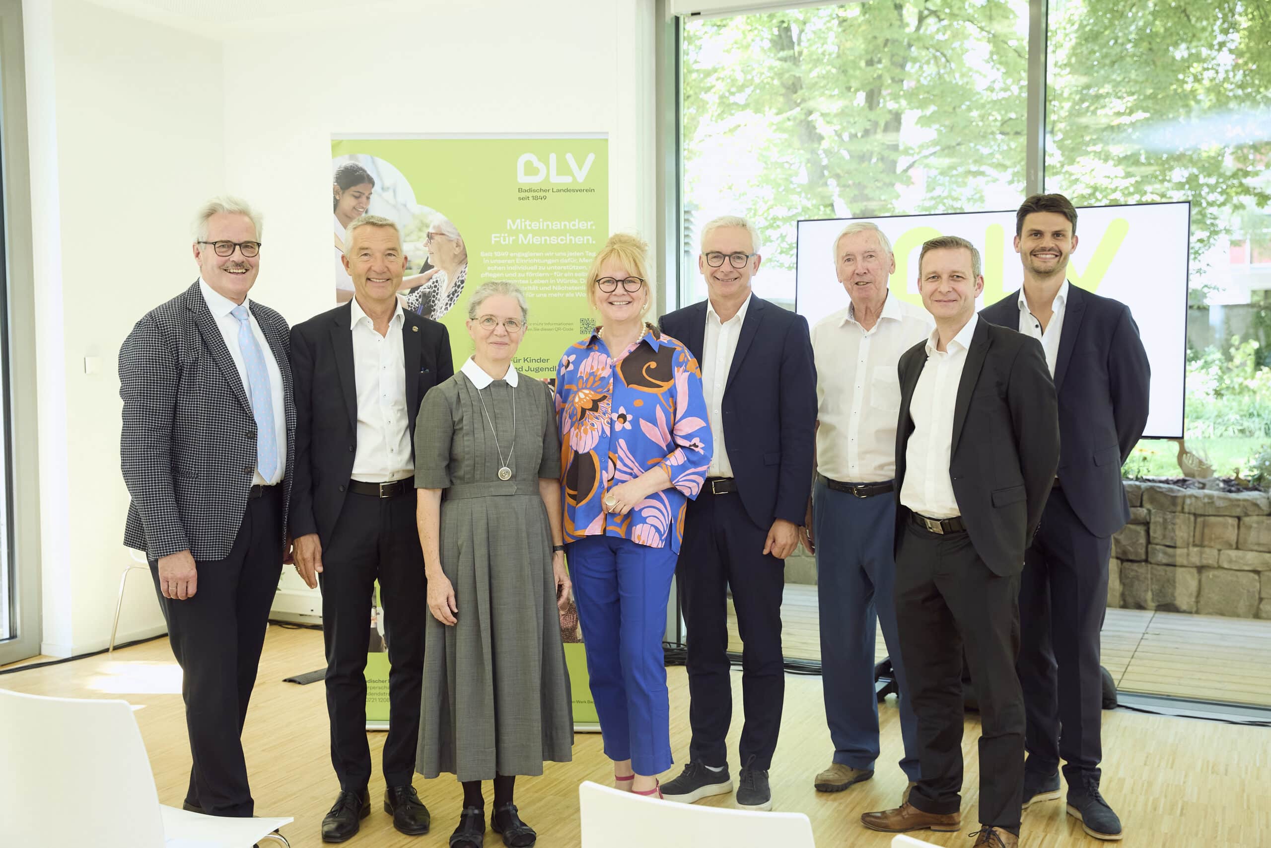 Eine Gruppe Menschen steht nebeneinander und lächelt in die Kamera. Sie tragen festliche Kleidung. Im Hintergrund sieht man einen Aufsteller des BLV und blickt durch große Fenster auf die Bäume im Garten des Friedensheims. Von links nach rechts sind zu sehen: Willy Kolarsch (Geschäftsführung GIG mbH), Dr. Martin Lenz (Bürgermeister Stadt Karlsruhe), Oberin Schwester Hildegund Fieg (Evangelisches Diakonissenhaus Bethlehem), Christine Jung-Weyand (Vorstand BLV), Dr. Christoph Schnaudigel (Landrat Landkreis Karlsruhe), Prof. Axel Göhringer (Verwaltungsratsvorsitzender BLV), Frank Derix (BWKG Baden-Württembergische Krankenhausgesellschaft e.V.), Pablo Müller Camarena (Geschäftsführung ACE Ingenieurconsult GmbH & Co. KG.)