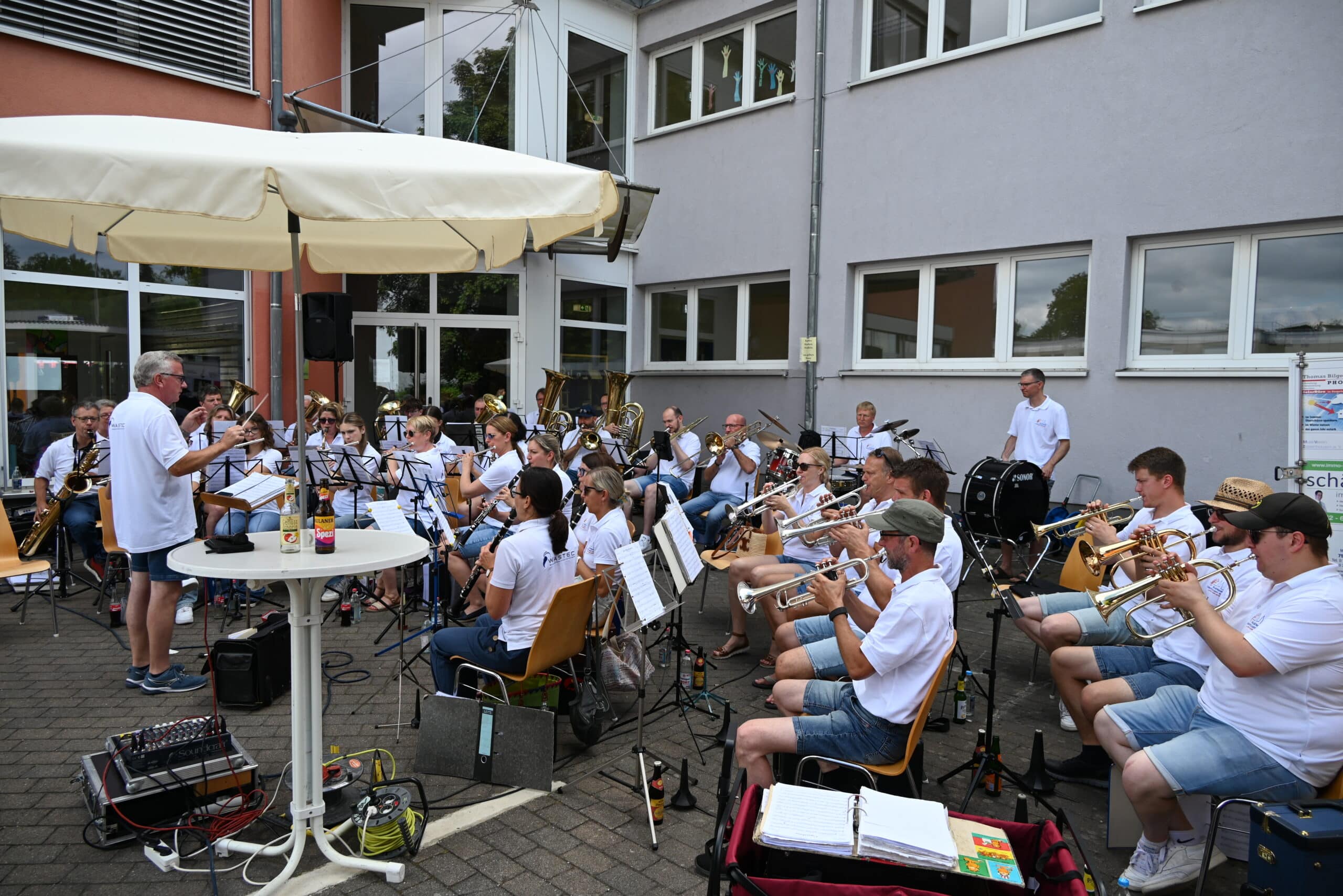 Quelle: BLV Die Musiker des Musikvereins Zaisenhausen sitzen auf Stühlen vor dem Hauptgebäude der Hohbergschule und spielen ihre Instrumente: Trompeten, Posaunen, Tuba, Klarinetten, Querflöte, Saxophon und viel mehr. Ein Mann steht vor dem Orchester und dirigiert. Alle tragen weiße Poloshirts.