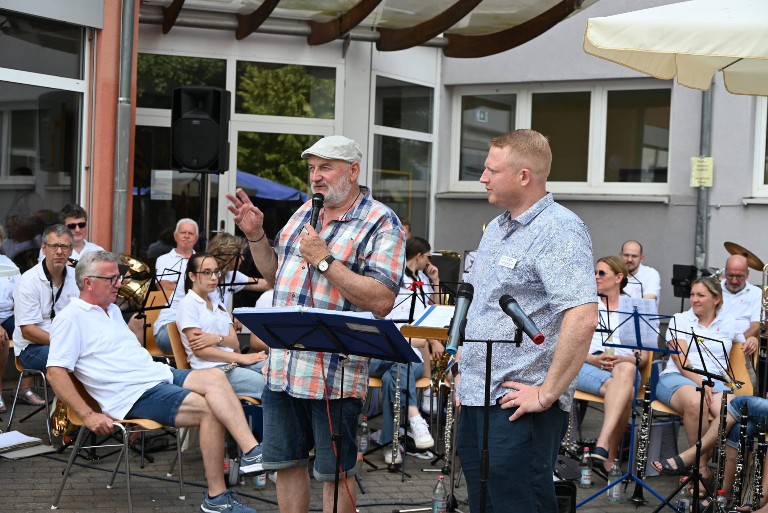 Quelle: BLV Peter Miebach steht neben Christoph Röckinger und hält eine Rede. Er trägt eine Schiebermütze und ein kariertes Hemd und hält ein Mikrophon in der Hand. Im Hintergrund sitzen die Mitglieder eines Orchesters. Sie haben ihre Musikinstrumente vor sich abgestellt und hören zu.