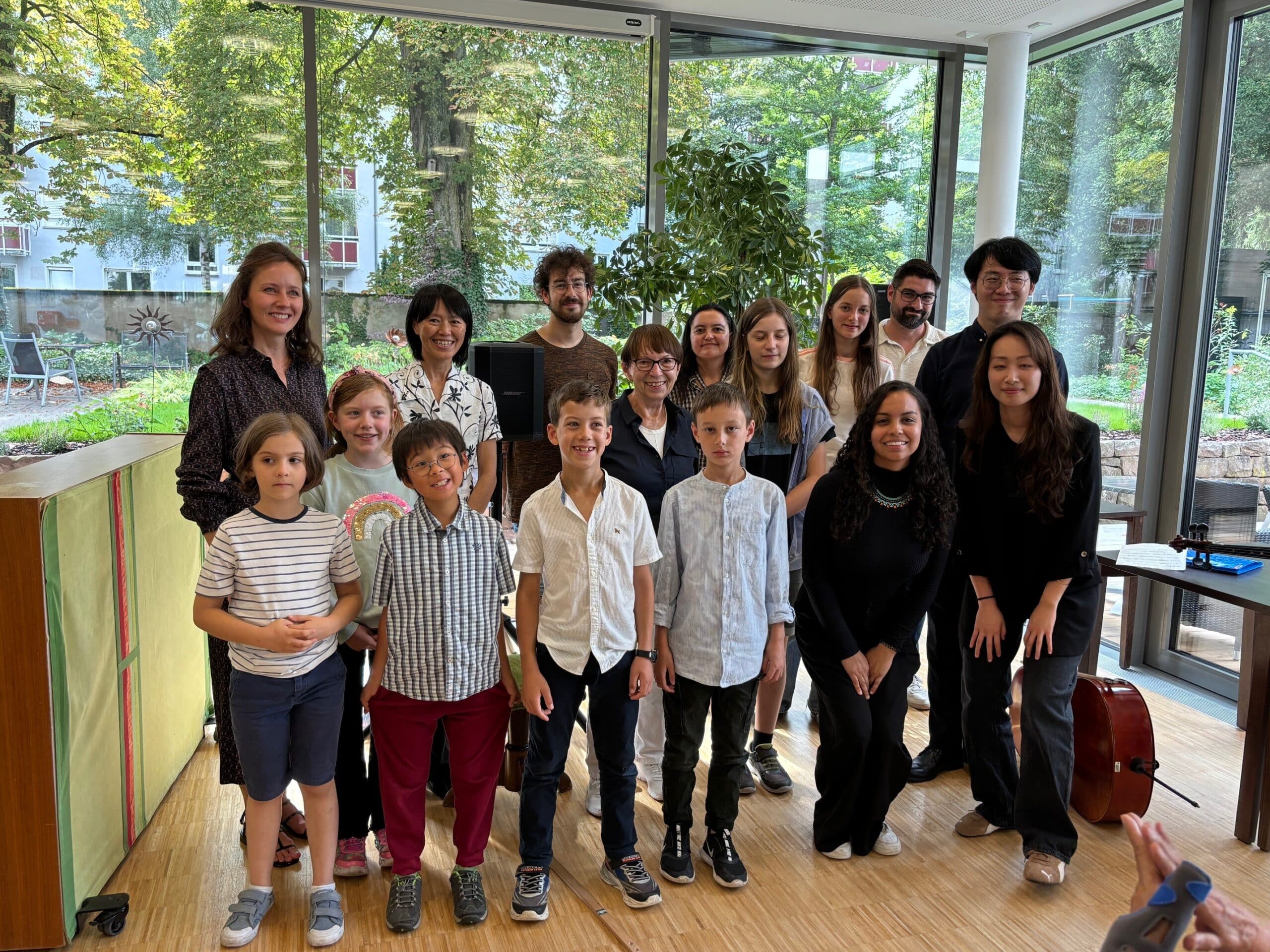 Mehrere Personen stehen nebeneinander und lächeln in die Kamera. Es sind vor allem junge Erwachsene und Kinder. Im Hintergrund sieht man den Bäume, Büsche und eine Wiese - der Garten des Friedensheims.