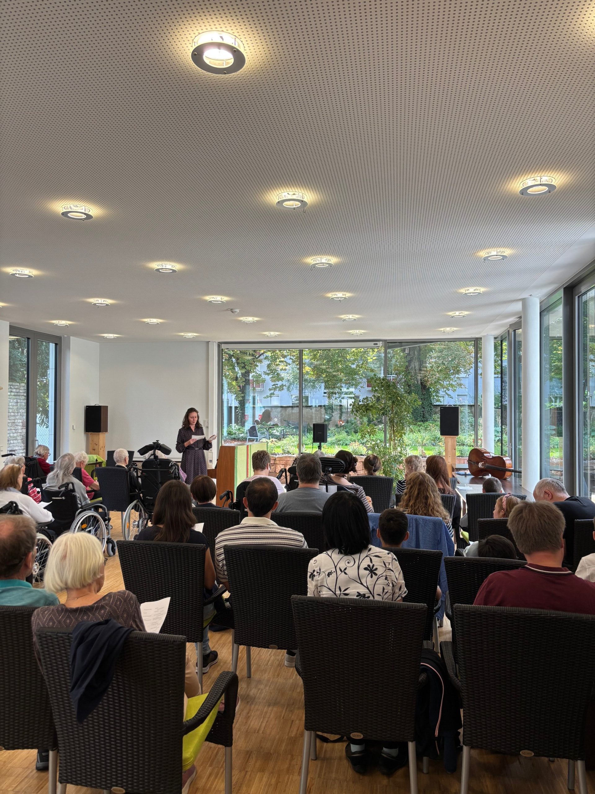 Eine Frau steht vor einem Publikum und liest etwas von einem Blatt ab. Einige der Menschen im Publikum sitzen im Rollstuhl.