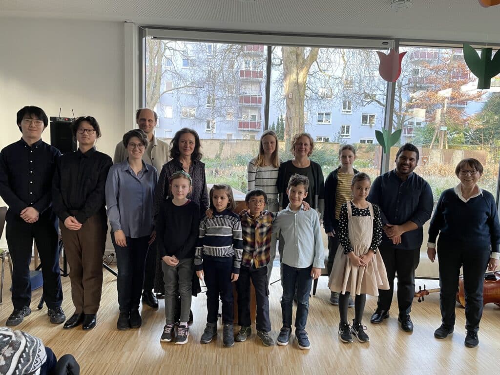 15 Personen stehen nebeneinander. Sie haben Hemd, Bluse oder Kleid an und lächeln. Durch die großen Scheiben im Hintergrund sieht man den herbstlichen Garten des Friedensheims mit seinen Büschen und Bäumen.