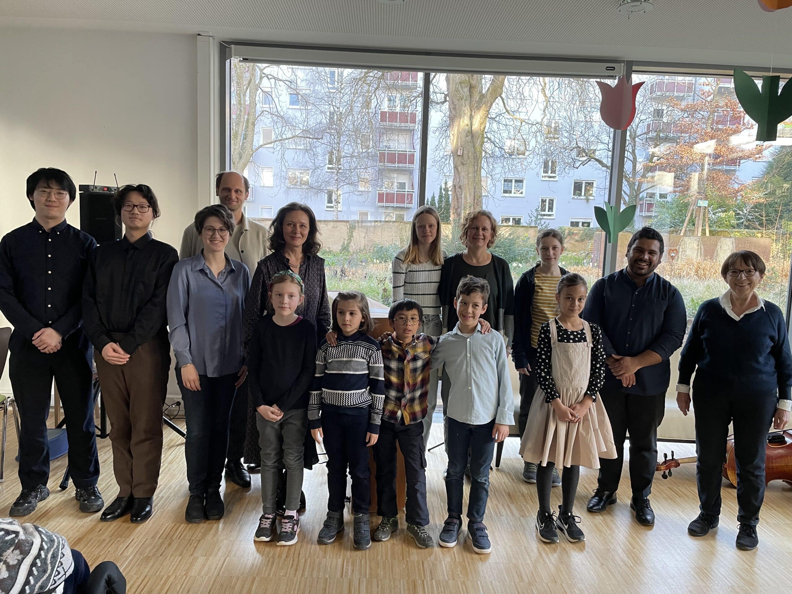 15 Personen stehen nebeneinander. Sie haben Hemd, Bluse oder Kleid an und lächeln. Durch die großen Scheiben im Hintergrund sieht man den herbstlichen Garten des Friedensheims mit seinen Büschen und Bäumen.