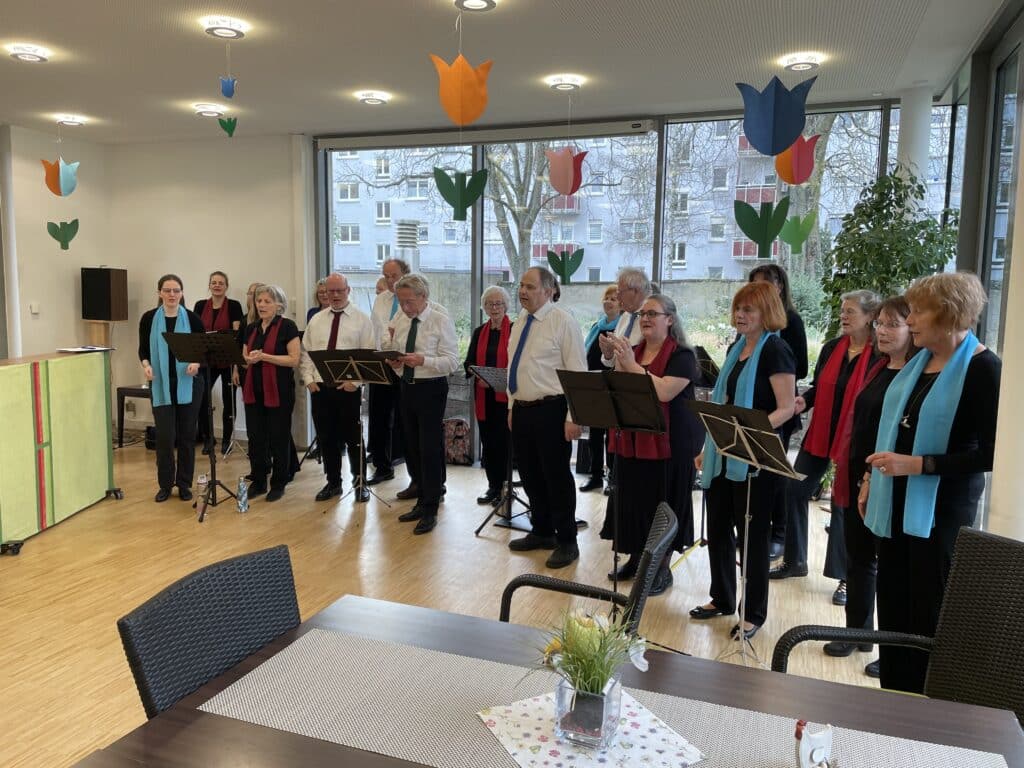 21 Personen stehen nebeneinander vor Notenständern und singen. Die Frauen tragen alle schwarze Shirts und ein hellblaues oder dunkelrotes Tuch. Die sechs Männer stehen in der Mitte der Gruppe und tragen weiße Hemden mit Krawatte. Auf den Notenständern liegen Textbücher, von denen die Personen ablesen. Durch die großen Scheiben im Hintergrund sieht man den Garten des Friedensheims mit seinen Büschen und Bäumen.