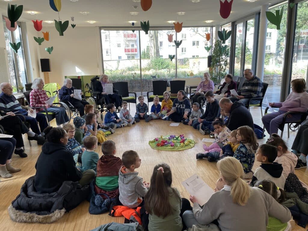 Viele Kinder sitzen im Kreis auf dem Fußboden um ein Tuch mit bunten Papierblumen. Drum herum stehen, ebenfalls im Kreis, Stühle, auf denen einige ältere Personen sitzen. Sie alle halten Papierblätter mit Text in den Händen. Durch die großen Scheiben im Hintergrund sieht man den Garten des Friedensheims mit seinen Büschen und Bäumen.