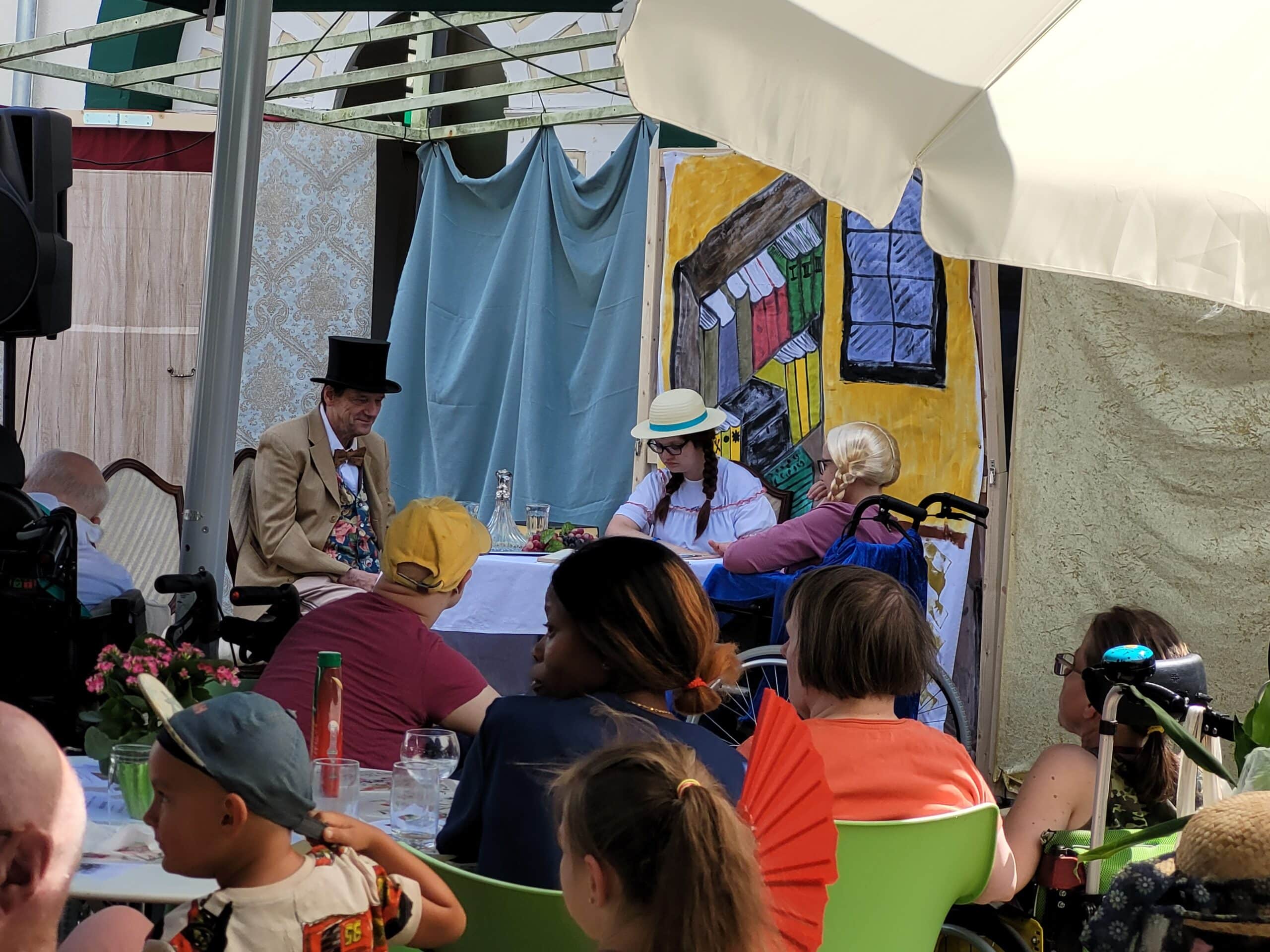 Zwei Frauen und ein Mann spielen auf der Bühne Theater. Der Mann trägt ein beiges Jackett, eine geblümte Weste, eine Fliege und einen schwarzen Zylinder. Eine der Frauen trägt eine weiße Bluse, schwarze, geflochtene Zöpfe und einen Strohhut. Die andere Frau sitzt in einem Rollstuhl und trägt ein dunkelrotes Shirt und eine blonde Perücke. Im Hintergrund ist ein Bücherregal auf eine Stoffbahn aufgemalt. Im Vordergrund sitzen mehrere Personen im Publikum und schauen zu.