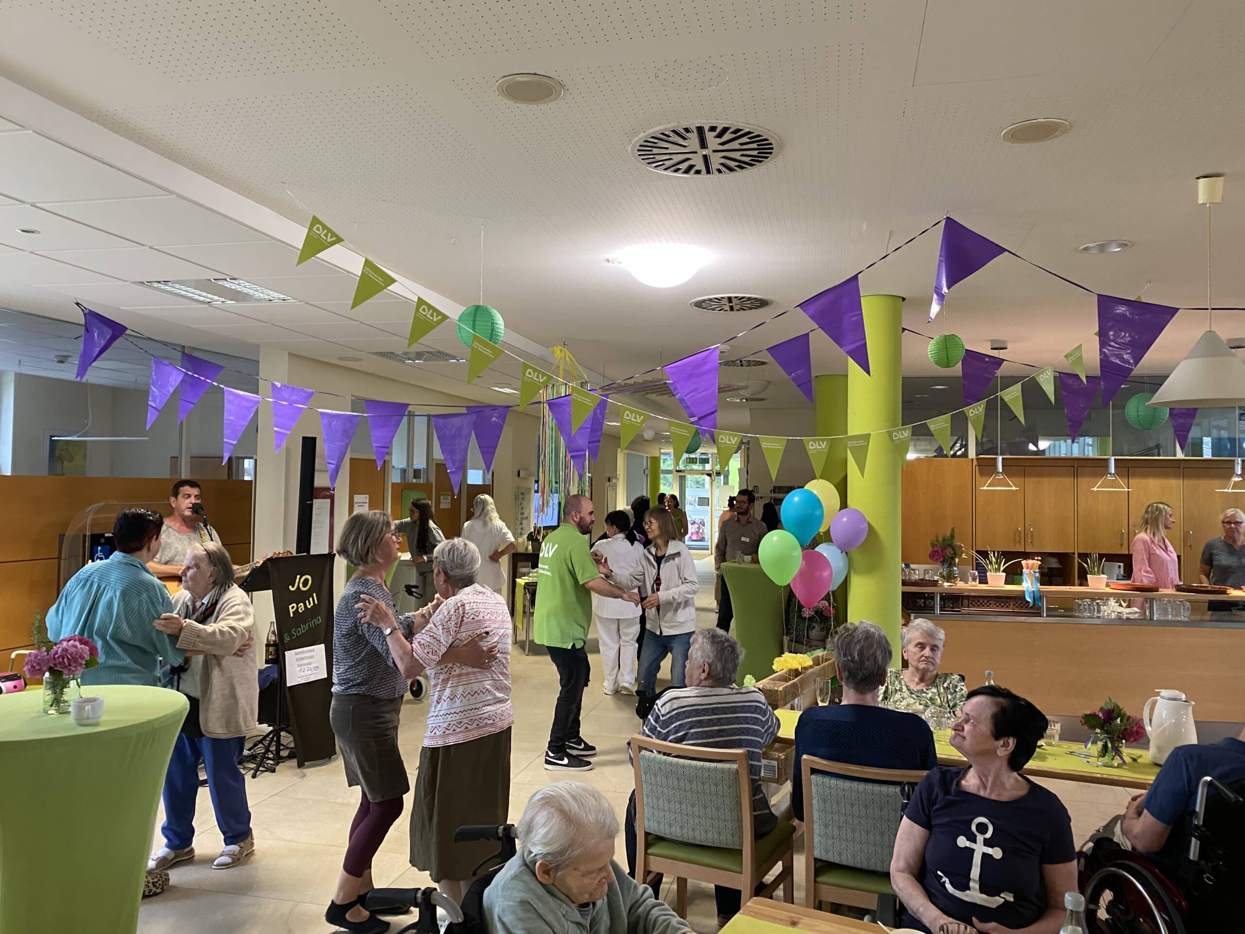 Mehrere ältere Personen beim Paartanz. Im Hintergrund spielt ein Musiker Gitarre und singt. Im Vordergrund sitzen weitere Menschen an geschmückten Tischen. Die Decke ist mit grünen und violetten Wimpelketten geschmückt.