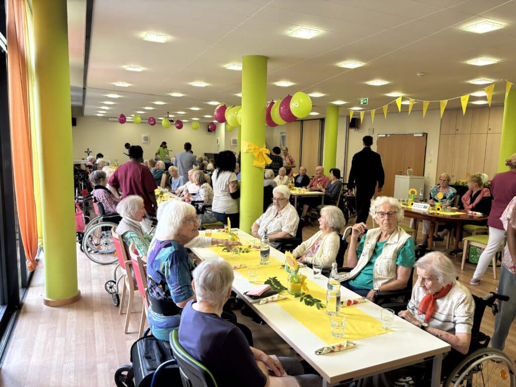 Ach ältere Frauen sitzen an einem langen Tisch. Im Hintergrund sieht man viele weitere Menschen, die an ähnlichen Tischen sitzen. Die Tische sind mit einem gelben Läufer, grünen Zweigen und gebastelten Sonnenblumen aus Papier geschmückt. An der Decke hängen Wimpelketten sowie grüne und violette Luftballons. Einige der Personen sitzen im Rollstuhl.