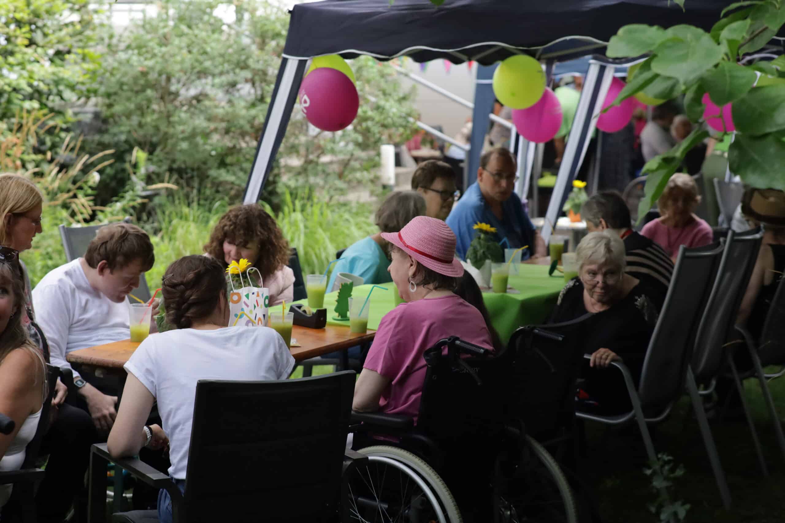 Zwölf Menschen sitzen unter einem Pavillon an Biertischen mit grüner Tischdecke. Der Pavillon ist mit grünen und violetten Luftballons geschmückt. Eine Person hat ein rosa T-shirt an und einen rosa Hut auf. Sie sitzt im Rollstuhl. Alle haben vor sich einen Plastikbecher mit einer grünen Flüssigkeit und einem Strohhalm stehen.