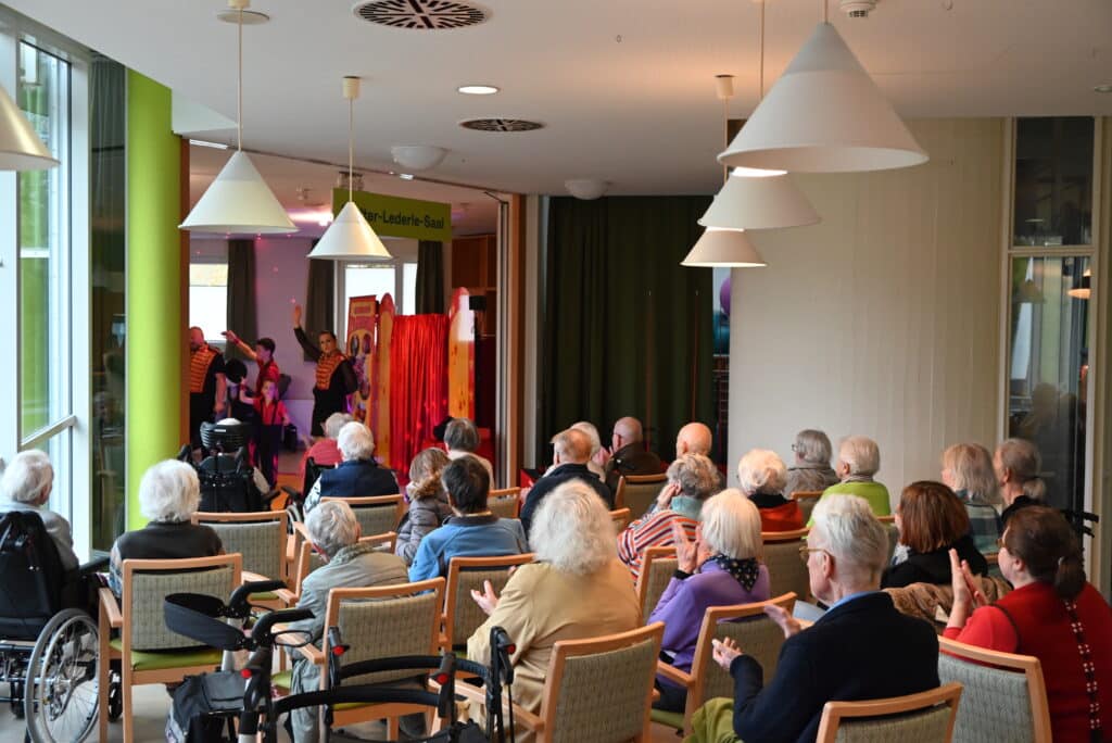 Eine große Gruppe älterer Menschen sitzt auf Holzstühlen und applaudiert. Zwei von ihnen sitzen im Rollstuhl. Auf der Bühne stehen zwei Erwachsene und zwei Kinder in rot-schwarzer Zirkuskleidung und winken in die Menge.