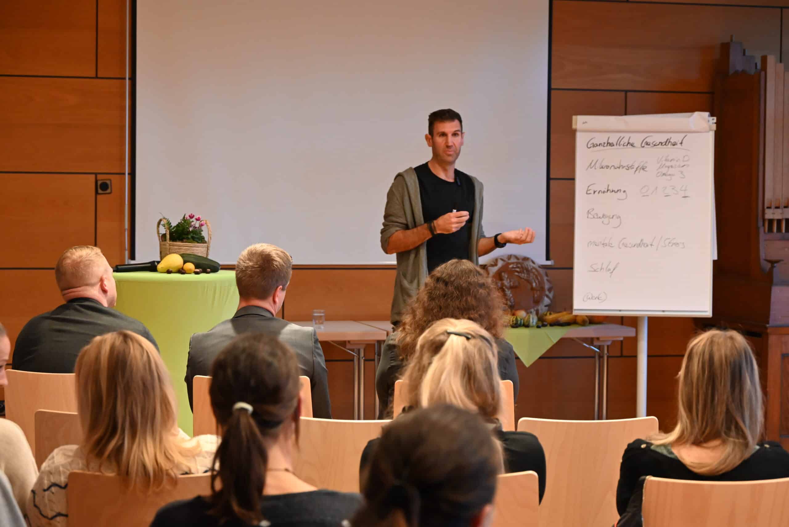 Mann steht vor einem Publikum und hält eine Rede. Neben ihm steht ein Board mit handschriftlichen Stichpunkten.