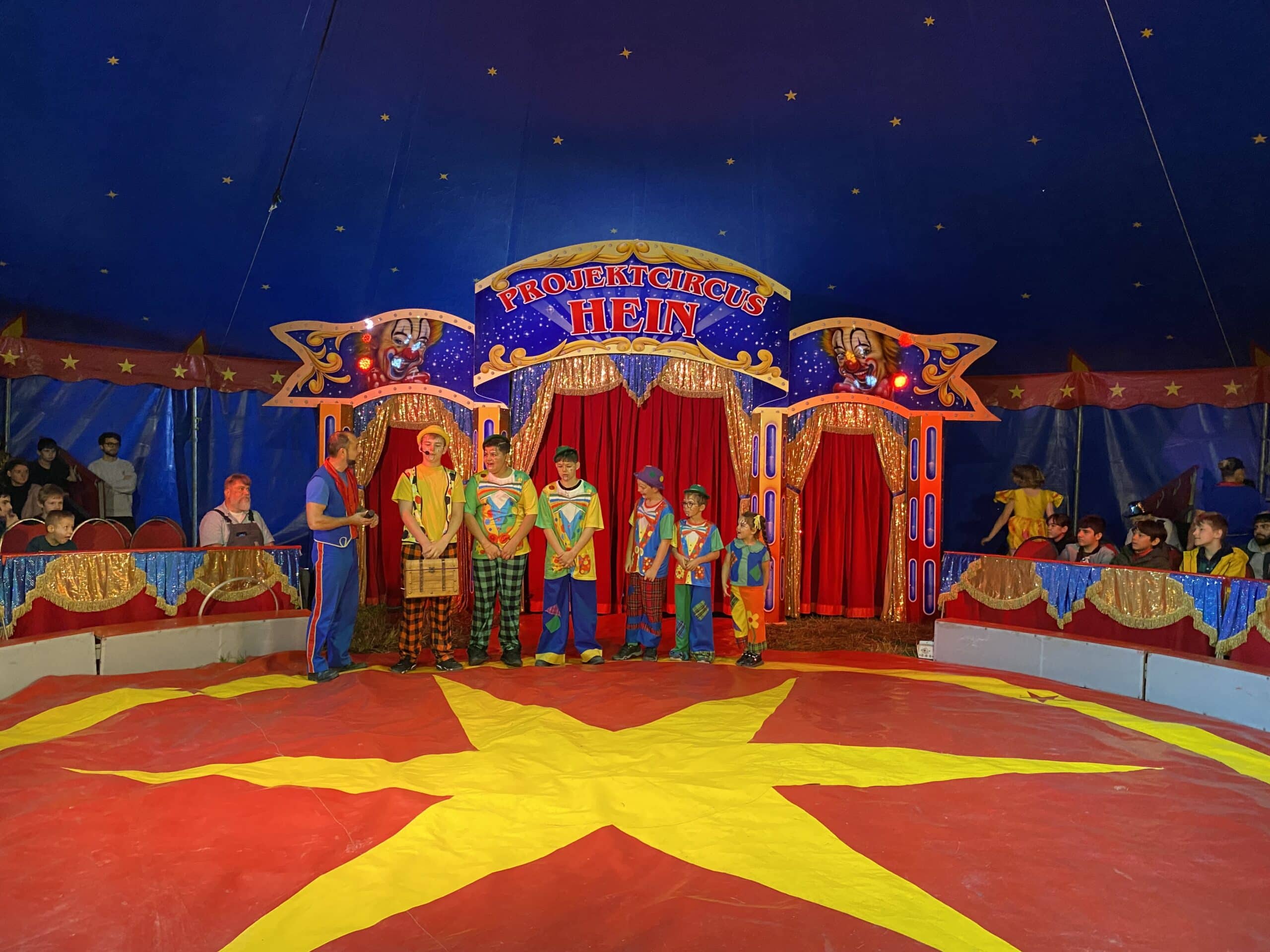 Sechs Kinder in bunten Clowns-Kostümen stehen am Rande der Zirkusmanege. Sind sind nach ihrer Größe aufgereiht. Der Zirkusdirektor spricht mit ihnen.