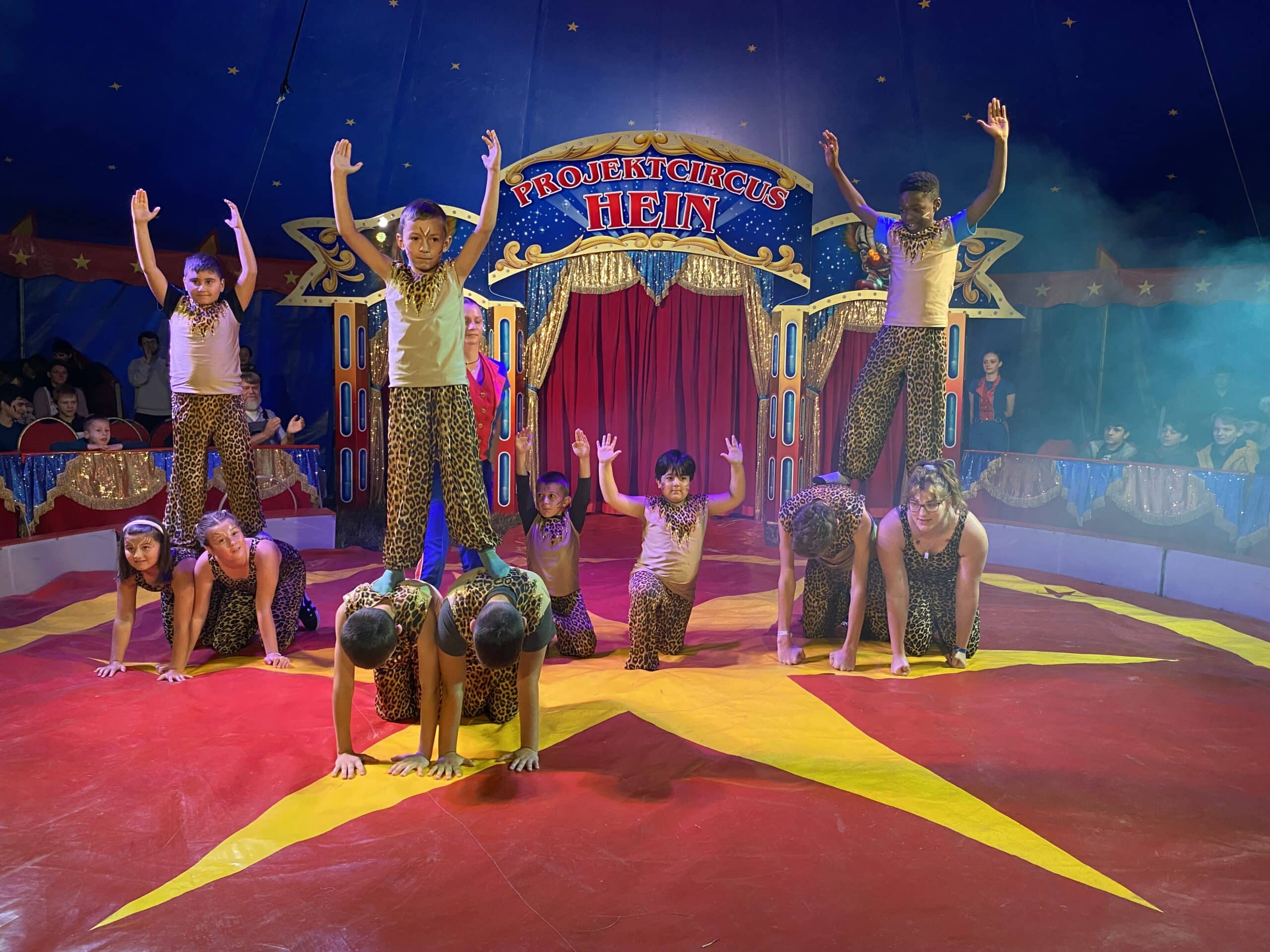 Quelle: BLV Mehrere Kinder in Kleidung mit Leopardenmuster zeigen eine Akrobatiknummer in drei Kleingruppen. Dabei knien jeweils zwei der Kinder, während ein drittes auf deren Rücken steht und die Arme in die Luft streckt.