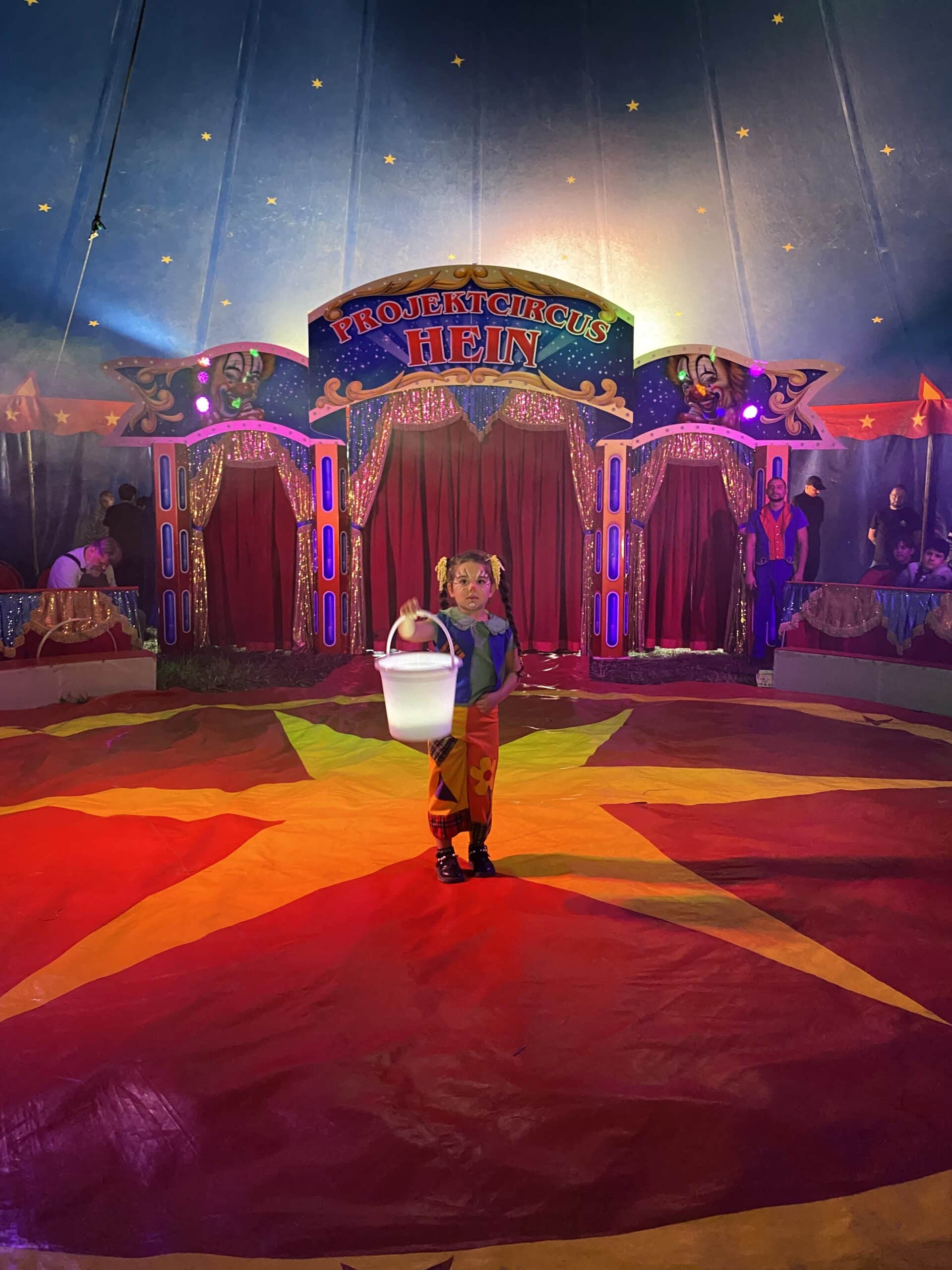 Quelle: BLV Ein kleines Mädchen steht in der Mitte einer Zirkusmanege. Sie ist geschminkt und trägt bunte Clowns-Kleidung. In der Hand hält sie einen großen weißen Eimer, den sie nach vorne streckt.