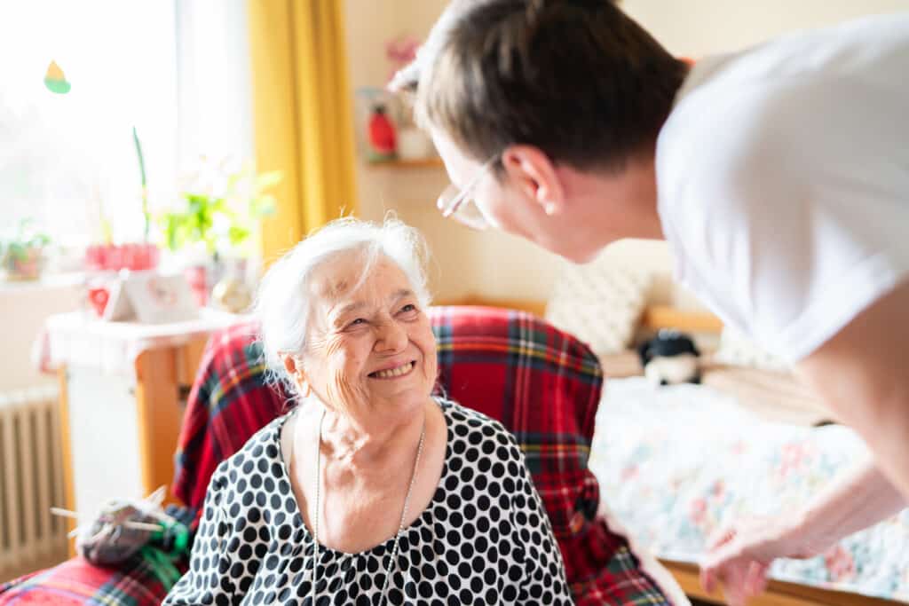 Ein Pfleger beugt sich zu einer älteren Frau hinunter. Diese lächelt zurück.