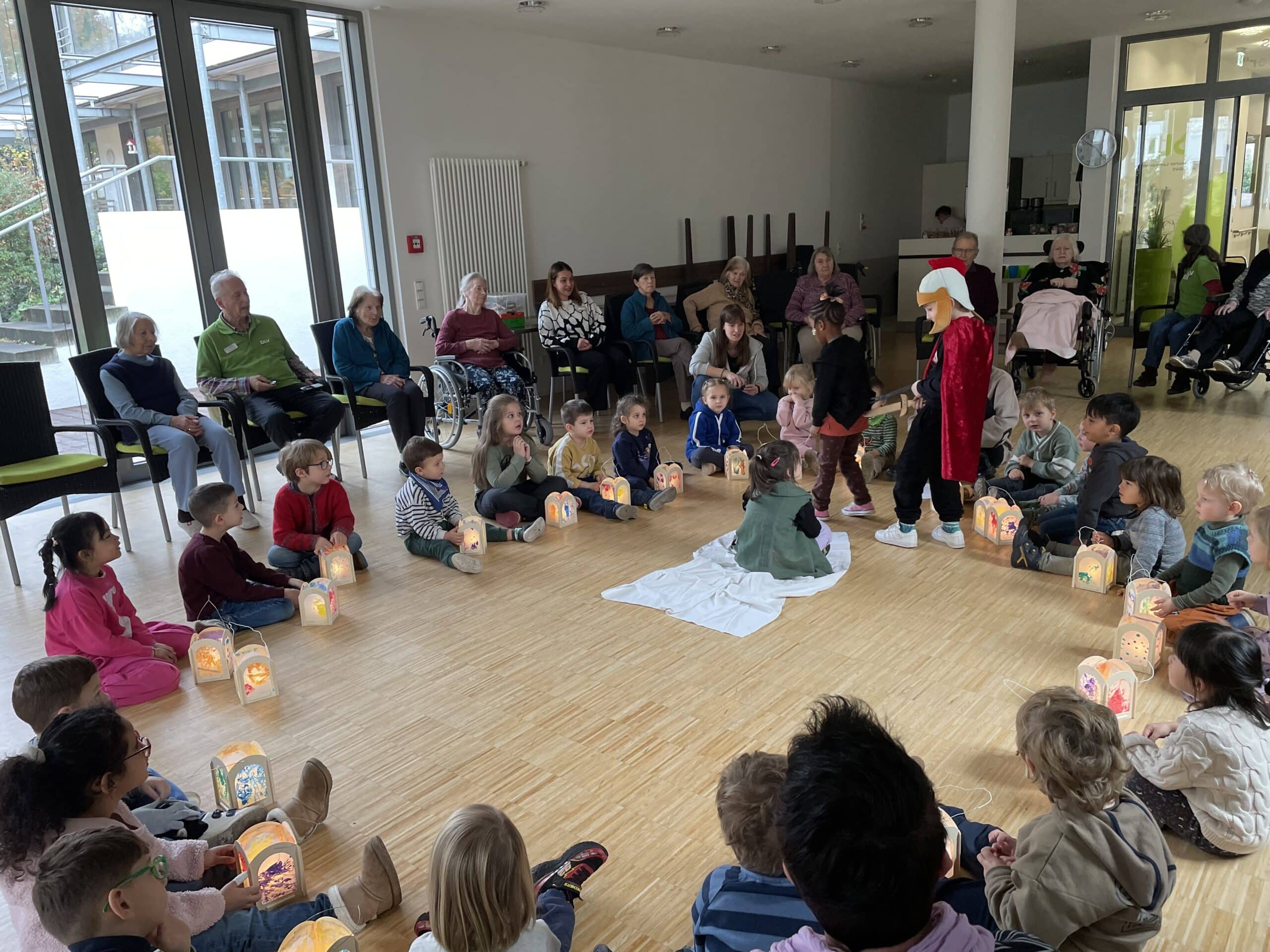 Kinder sitzen im Kreis auf dem Boden. Vor ihnen stehen Laternen auf dem Boden. Auf Stühlen sitzen ältere Menschen ebenfalls im Kreis um die Kinder herum.