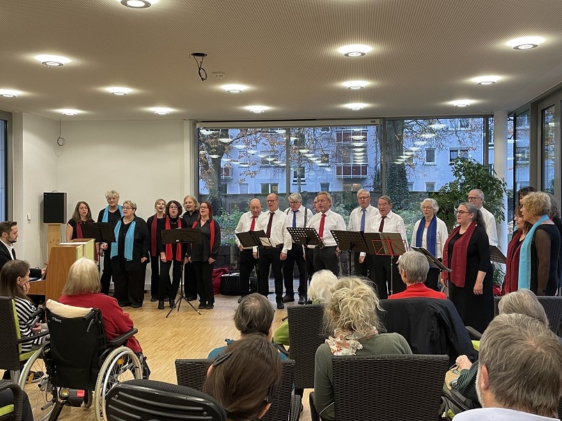 Etwa 20 Männer und Frauen stehen im Halbkreis und singen. Neben ihnen steht ein Klavier, auf dem ein Mann spielt. Vor ihnen sitzen mehrere ältere Menschen im Publikum und hören zu. Eine von ihnen sitzt in einem Rollstuhl