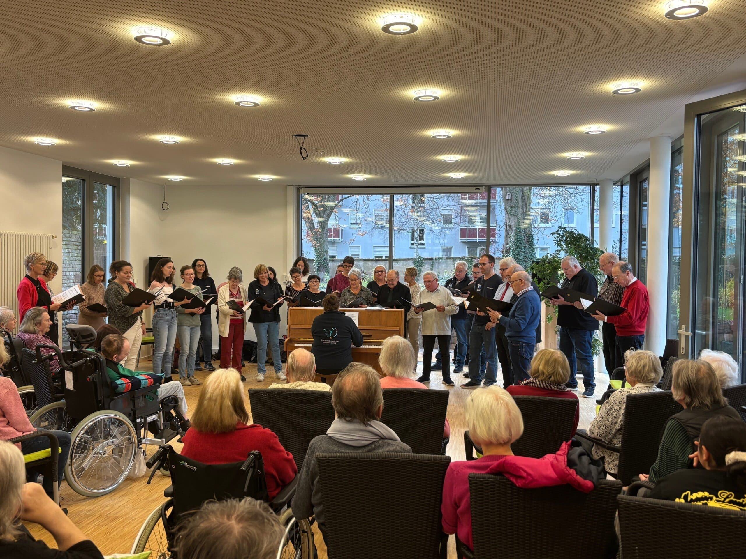 Mehrere Menschen stehen im Halbkreis um ein Klavier herum und singen. Auf dem Klavier spielt eine Frau. Vor dem Chor sitzen viele Menschen im Publikum auf Stühlen und höhren zu.