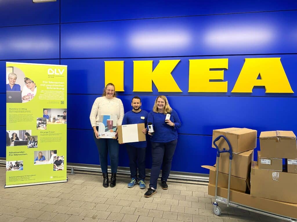 Drei Personen stehen vor einer blauen Wand. Darauf steht in großen, gelben Buchstaben IKEA geschrieben. Neben ihnen steht ein Wagen voller Pakete, auf der anderen Seite ein grüner Aufsteller mit BLV-Logo.