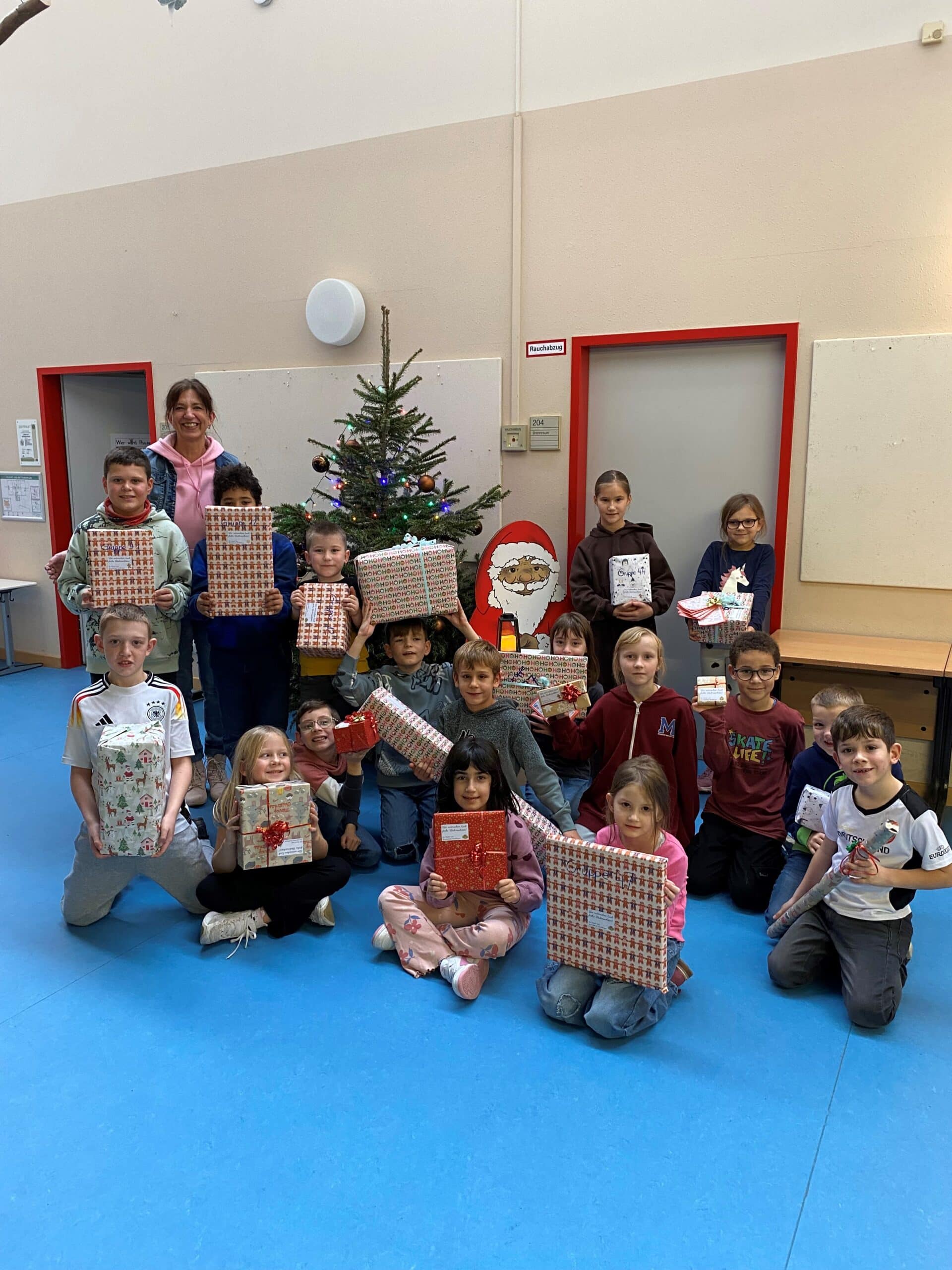 14 Kinder und eine Lehrerin stehen nebeneinander. Sie halten Weihnachtsgeschenke in ihren Händen.