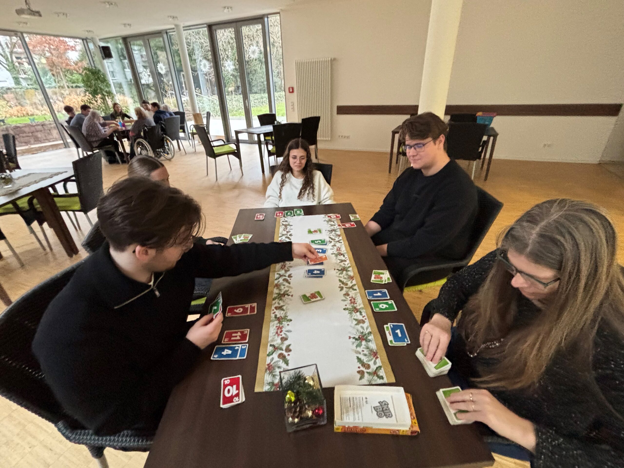 Drei junge Menschen sitzen mit zwei älteren Menschen am Tisch und spielen ein Kartenspiel. Im Hintergrund sieht man an einem anderen Tisch eine weitere Spielrunde.