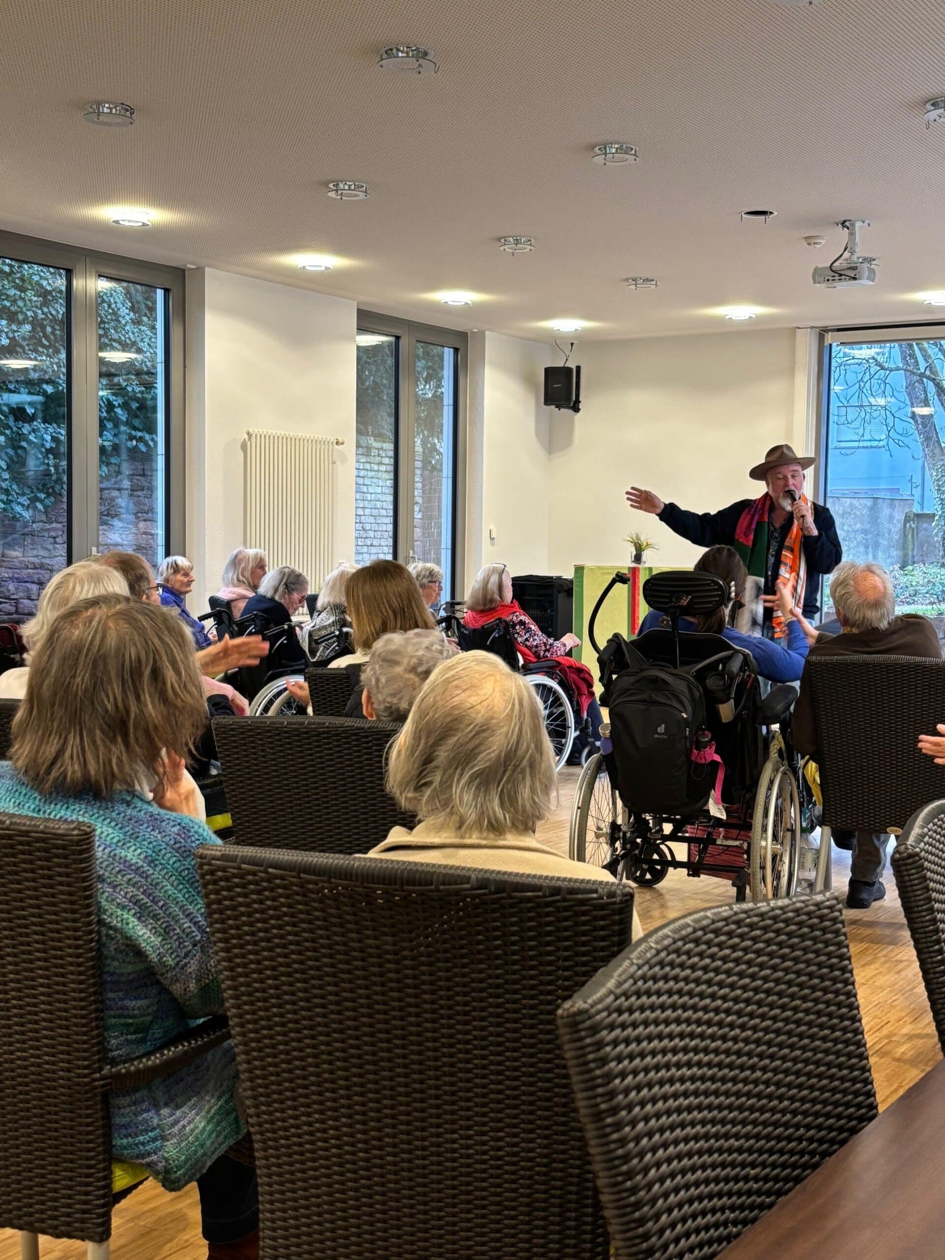Ein Mann mit Hut singt. Mehrere ältere Menschen sitzen auf Stühlen und höhren zu.
