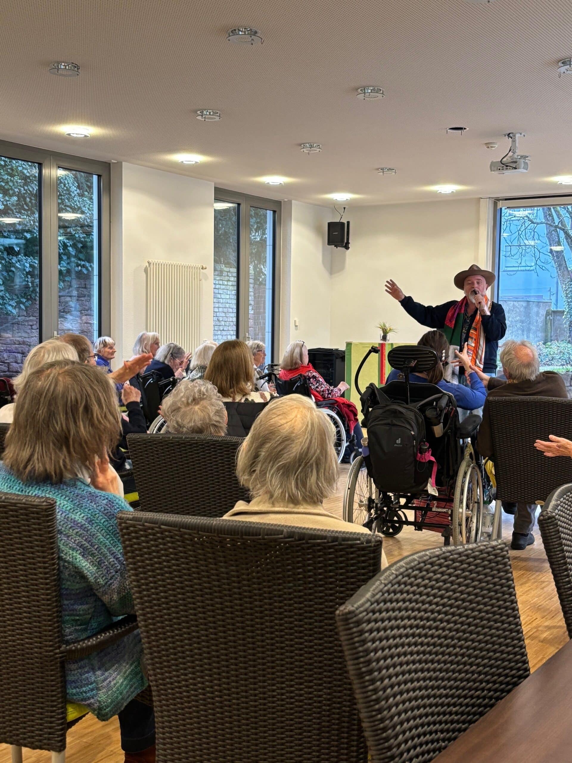 Ein Mann mit Hut singt. Mehrere ältere Menschen sitzen auf Stühlen und höhren zu.