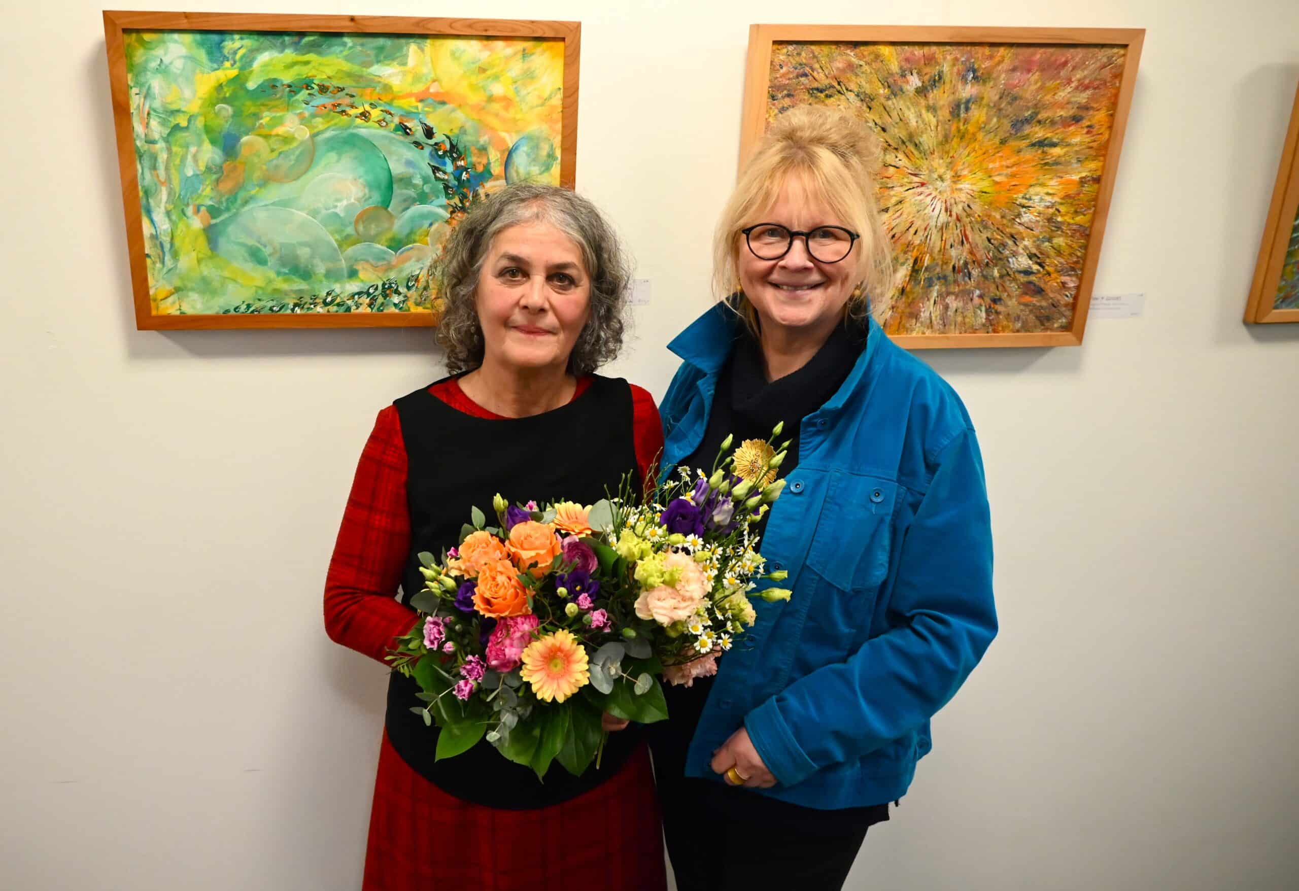 Die Künstlerin Helga Betsarkis und BLV-Vorstand Christine Jung-Weyand stehen nebeneinander vor den Kunstwerken, die Künstlerin hält einen bunten Blumenstrauß in den Händen.