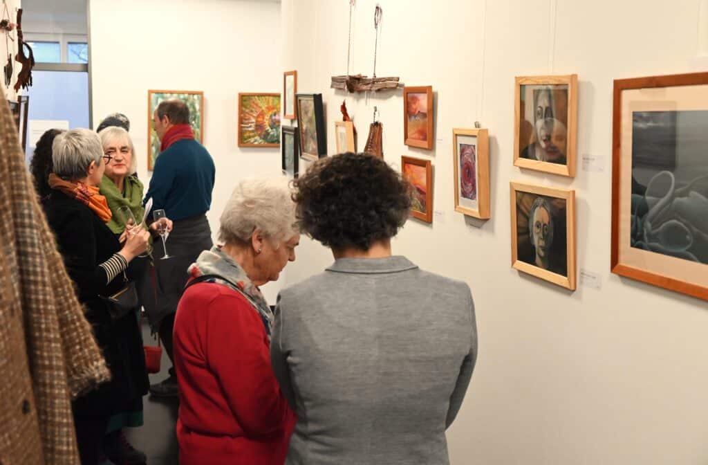 Einige Besucherinnen und Besucher betrachten die Kunstwerke von Helga Betsarkis.