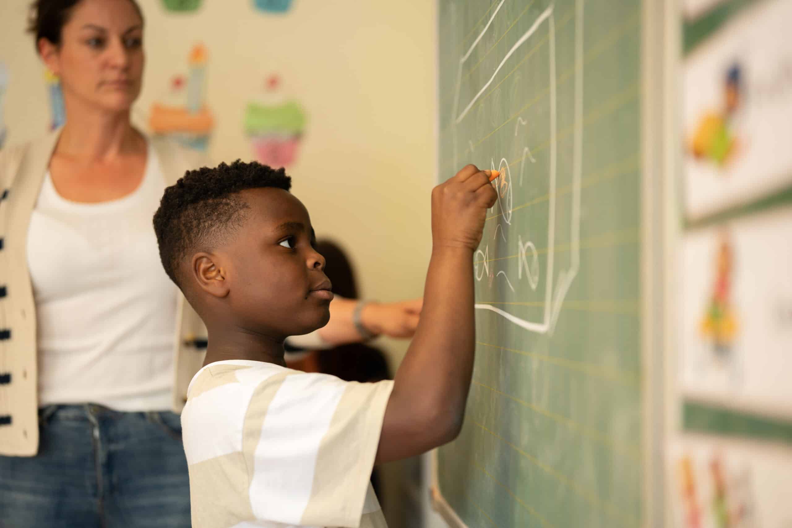 Quelle: BLV Ein Junge schreibt mit Kreide an die Tafel. Im Hintergrund steht eine Lehrerin und beobachtet ihn dabei.