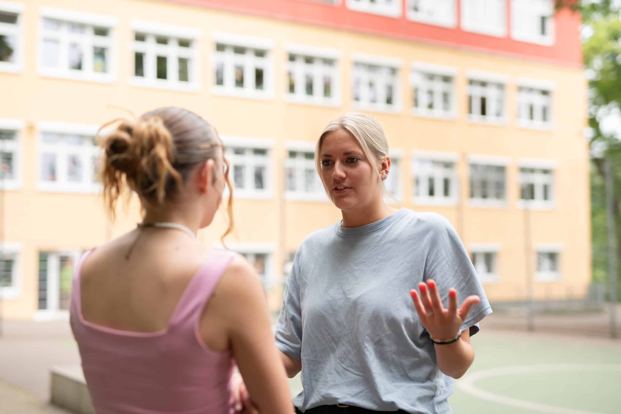 eine erwachsene Frau und ein Mädchen sprechen miteinander. Im Hintergrund ist die Schule Enzberg zu sehen.