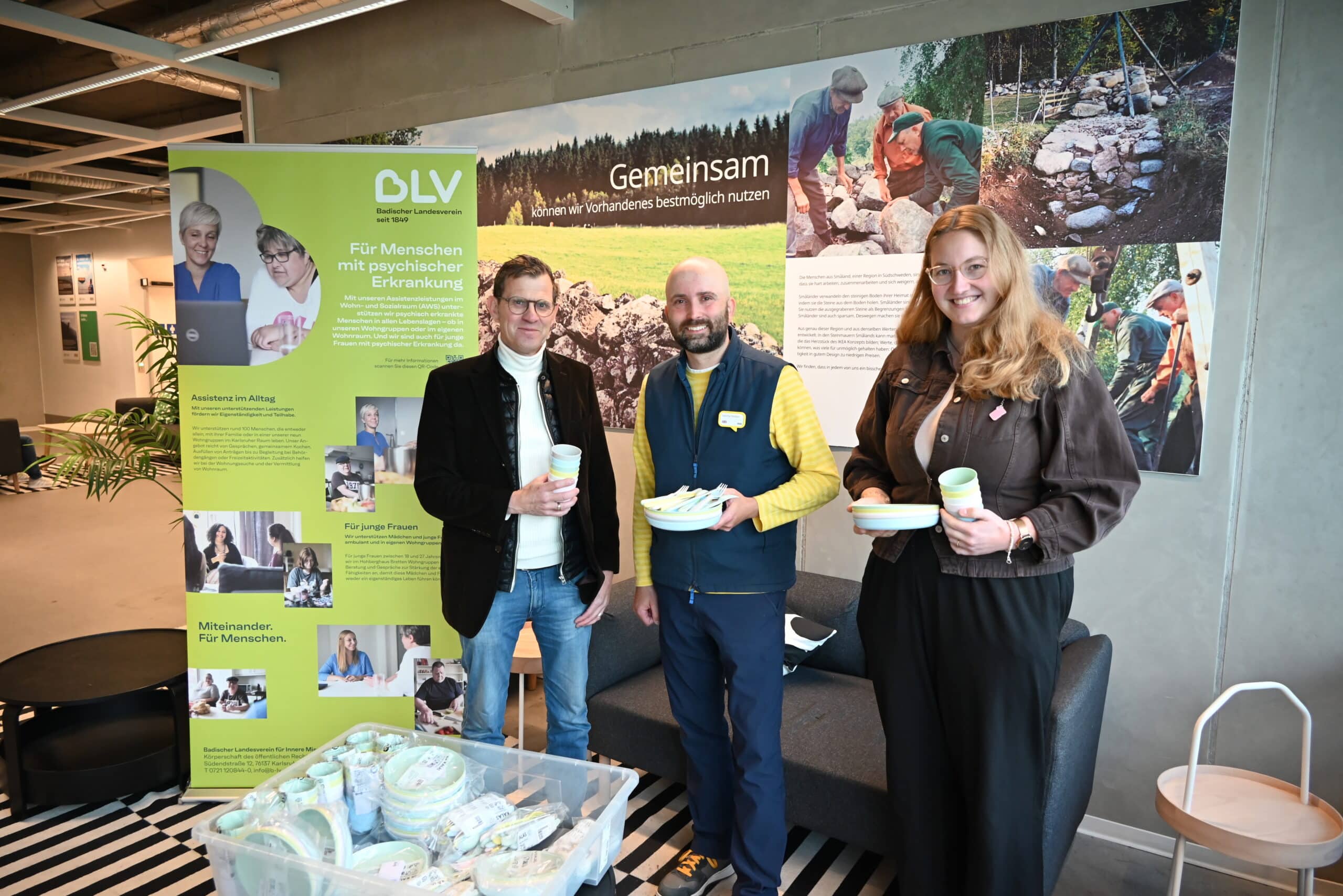 Zwei Männer und eine Frau stehen vor einem großen Plakat von IKEA auf dem steht "Gemeinsam können wir Vorhandenes bestmöglich nutzen" und einem hellgrünen Roll-Up auf dem steht "Für Menschen mit psychischen Erkrankungen", halten Plastikgeschirr in den Händen und lächeln in die Kamera.