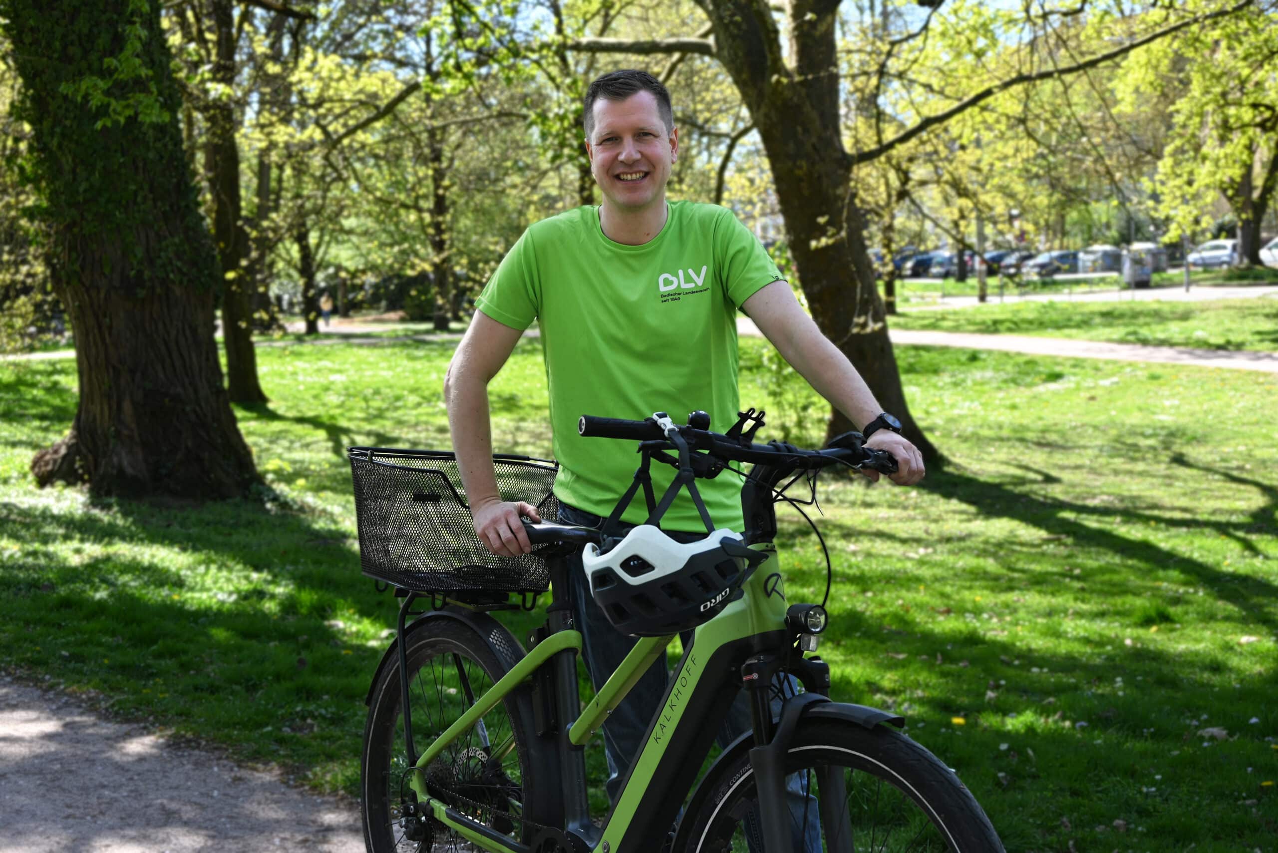 Timo Müller steht an seinem grünen Fahrrad vor Bäumen auf einer Wiese und lächelt in die Kamera.
