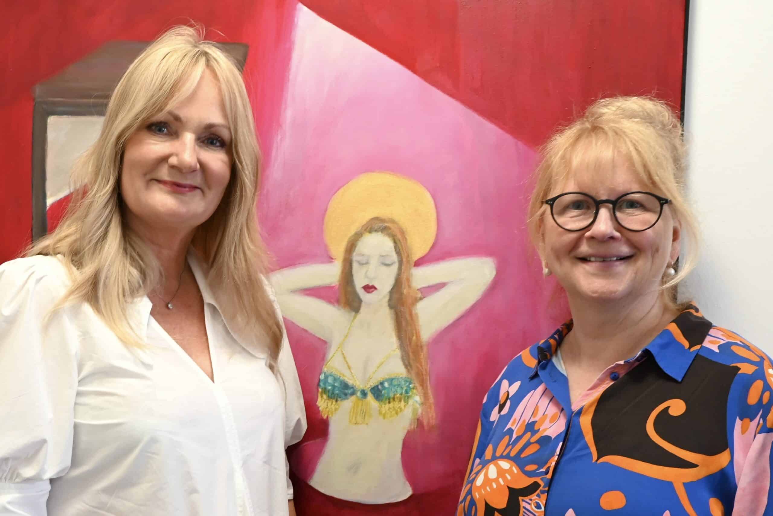Katharina Beckmann und Christine Jung-Weyand stehen vor einem Selbstporträt der Künstlerin Ariane Schmider und lächeln in die Kamera.