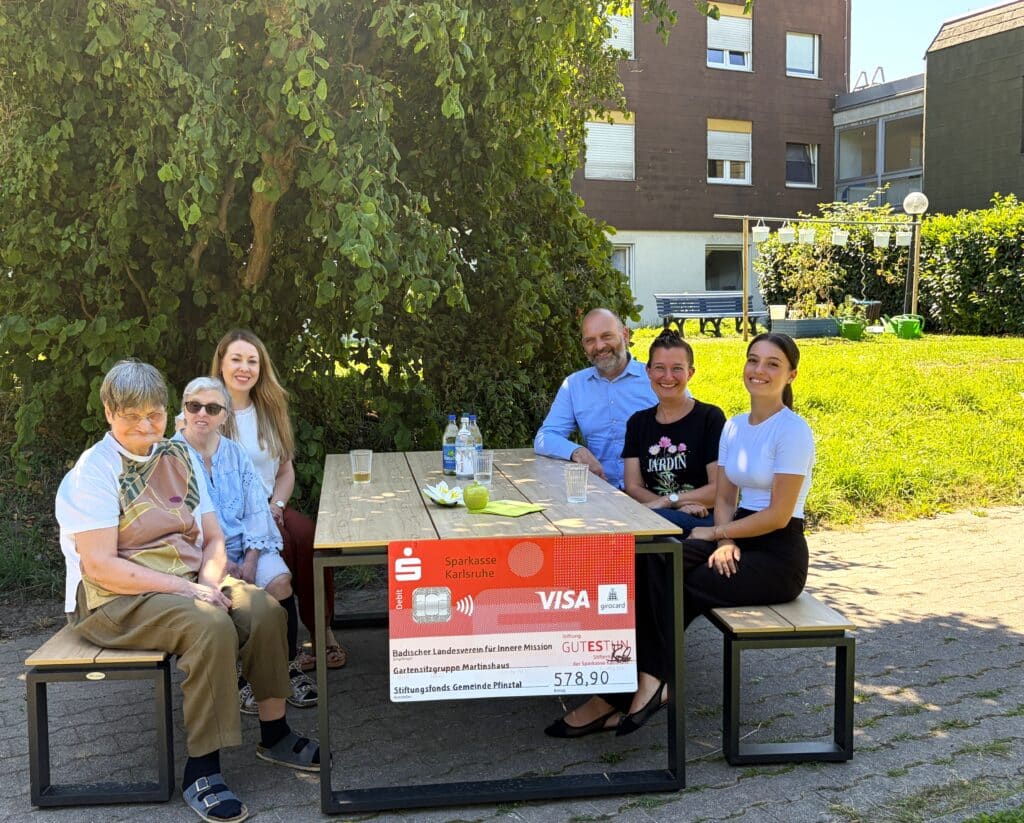 Bewohnerinnen des Martinshauses sitzen gemeinsam mit Evelyn Schieber, Einrichtungsleitung, Rainer Schuhmacher, Generationen- und Stiftungsberatung der Sparkassenstiftung GUTES TUN, Bürgermeisterin Nicola Bodner und Amelia Marrone, Mitarbeiterin der Sparkasse Karlsruhe auf den neuen Gartenmöbel.