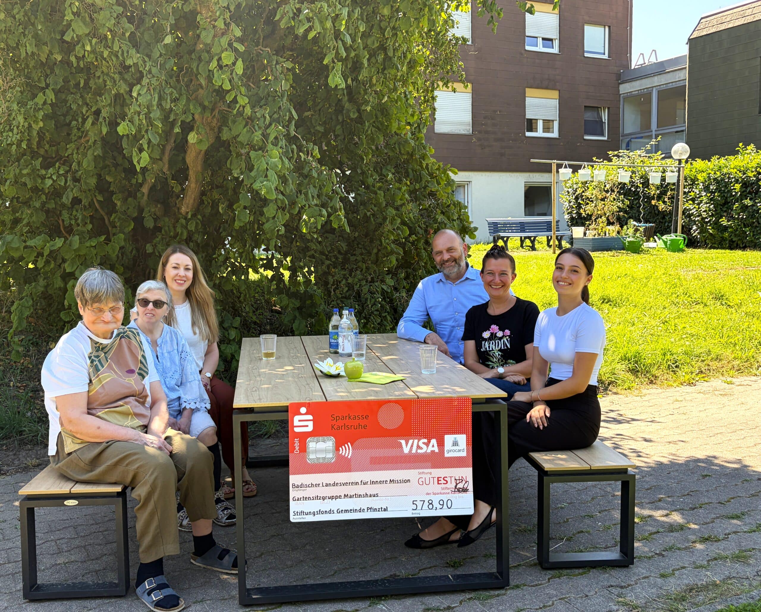 Bewohnerinnen des Martinshauses sitzen gemeinsam mit Evelyn Schieber, Einrichtungsleitung, Rainer Schuhmacher, Generationen- und Stiftungsberatung der Sparkassenstiftung GUTES TUN, Bürgermeisterin Nicola Bodner und Amelia Marrone, Mitarbeiterin der Sparkasse Karlsruhe auf den neuen Gartenmöbel.