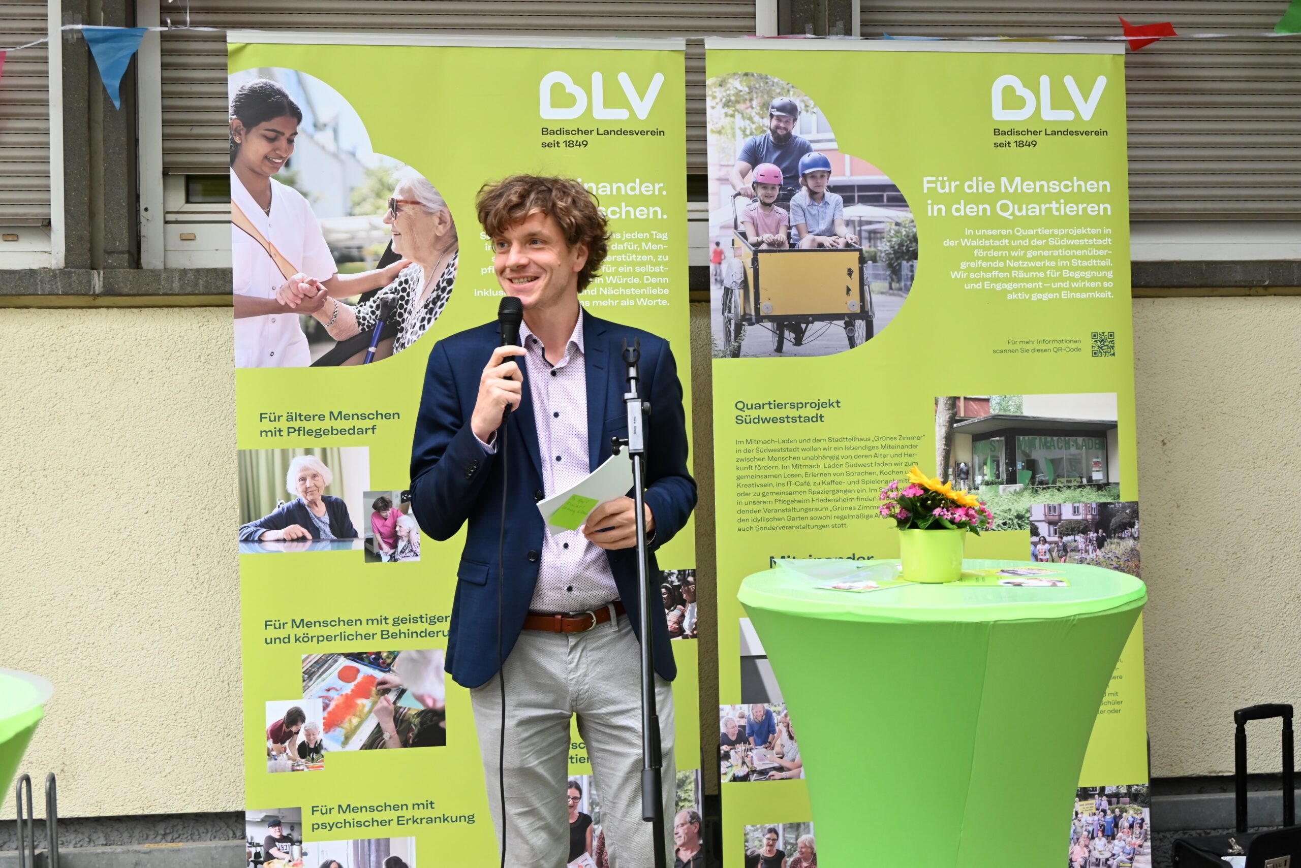 Quelle: BLV Dr. Raimund Bauer, Leiter der Strategischen Angebotsentwicklung des BLV steht vor zwei grünen Roll-Ups und spricht in ein Mikrofon. Neben ihm steht ein Stehtisch mit grüner Husse und einem Blumengesteck aus Sonnenblumen.