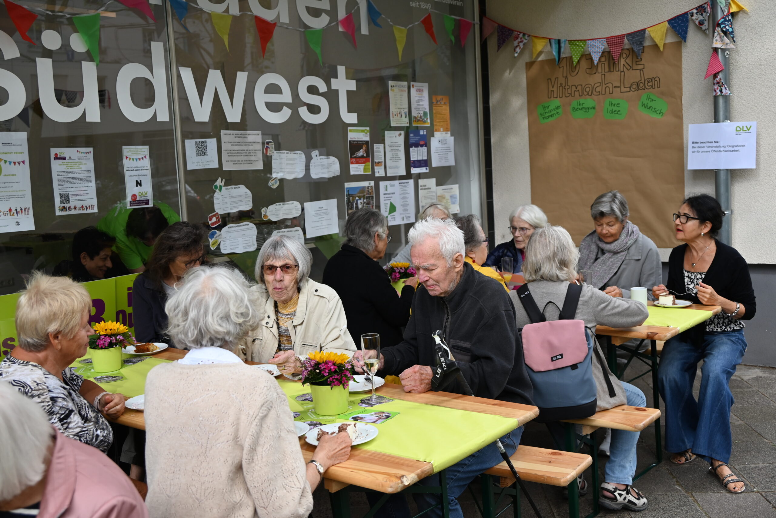Quelle: BLV Viele Menschen sitzen vor dem Mitmach-Laden Südwest auf Bierbänken und essen Kuchen. Auf den Tischen stehen bunte Blumensträuße in hellgrünen Töpfen. Im Hintergrund hängt eine bunte Girlande.