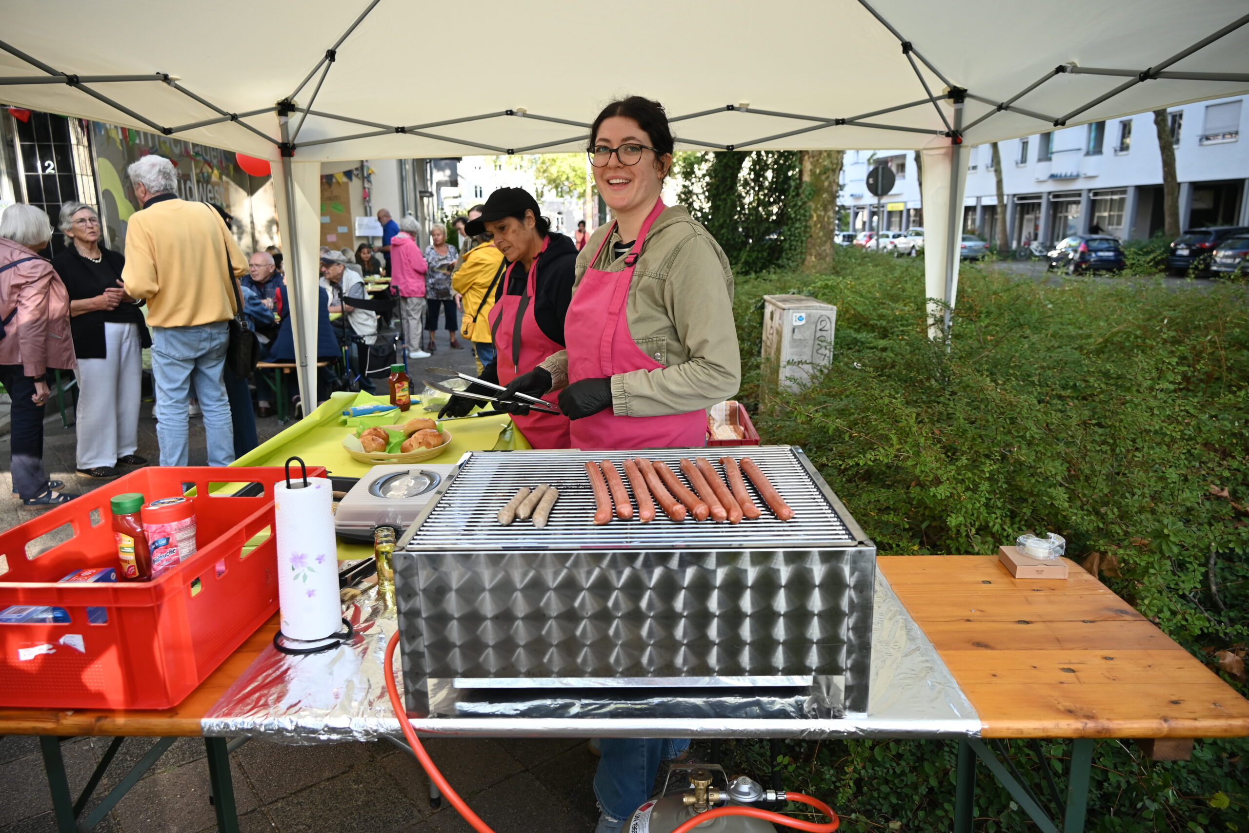 Quelle: BLV Zwei Frauen stehen am Grill und grillen Bratwürste. Sie tragen rosa Schürzen und lächeln in die Kamera. Im Hintergrund unterhalten sich mehrere Menschen.